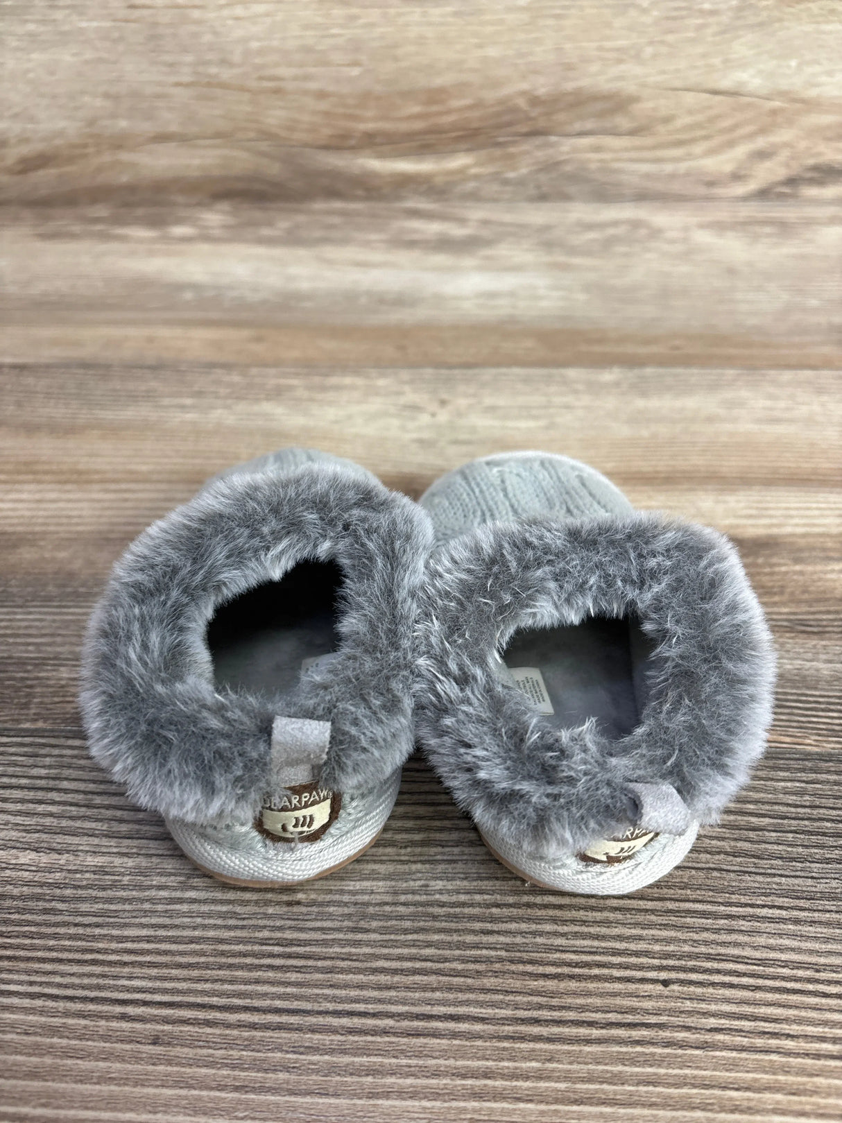 Gray faux fur lined baby slippers with Bearpaw label on wooden surface