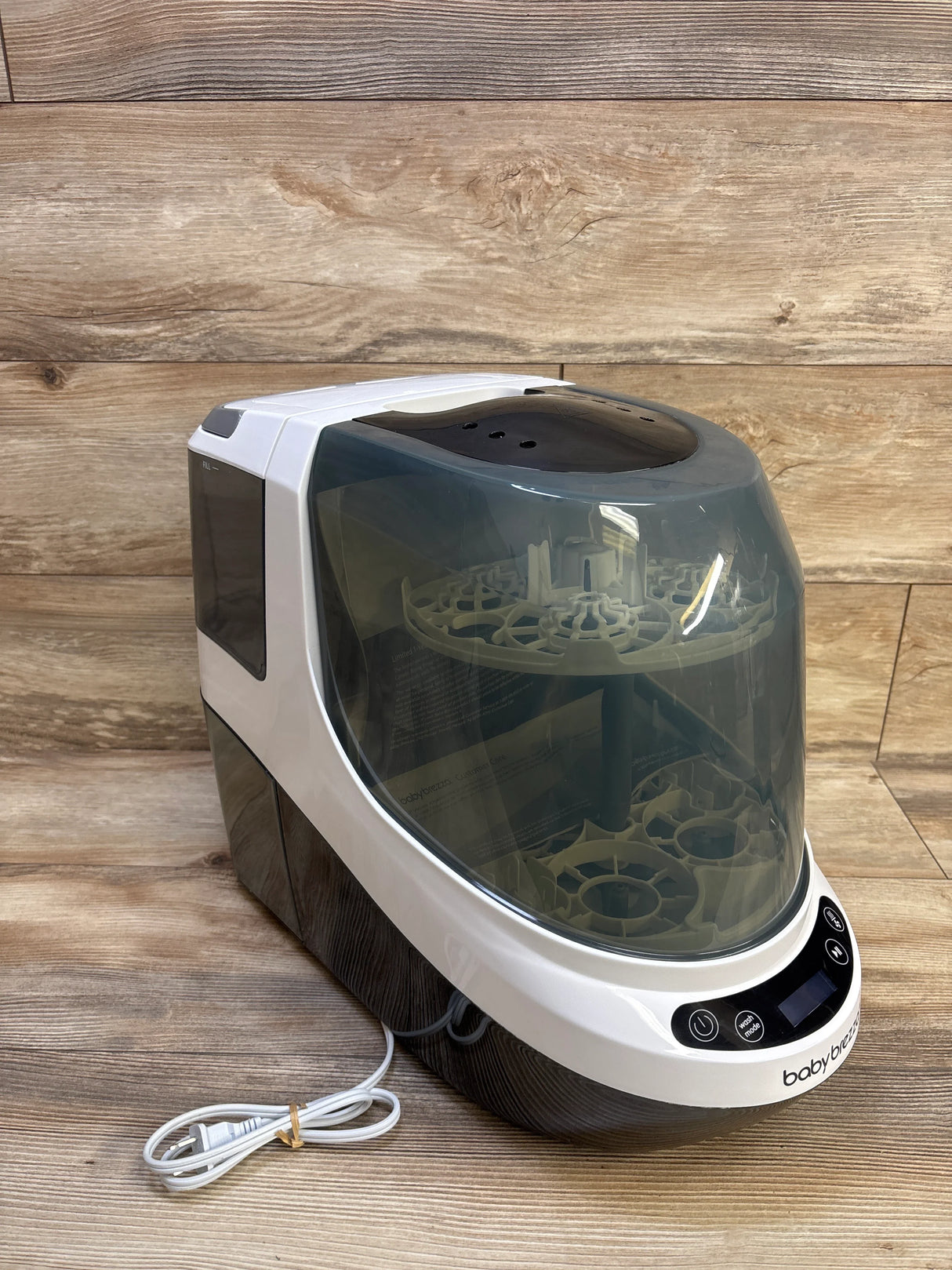Three-quarter view of Baby Brezza Bottle Washer Pro showcasing two-tier bottle racks and translucent dark lid with coiled power cord on wood backdrop