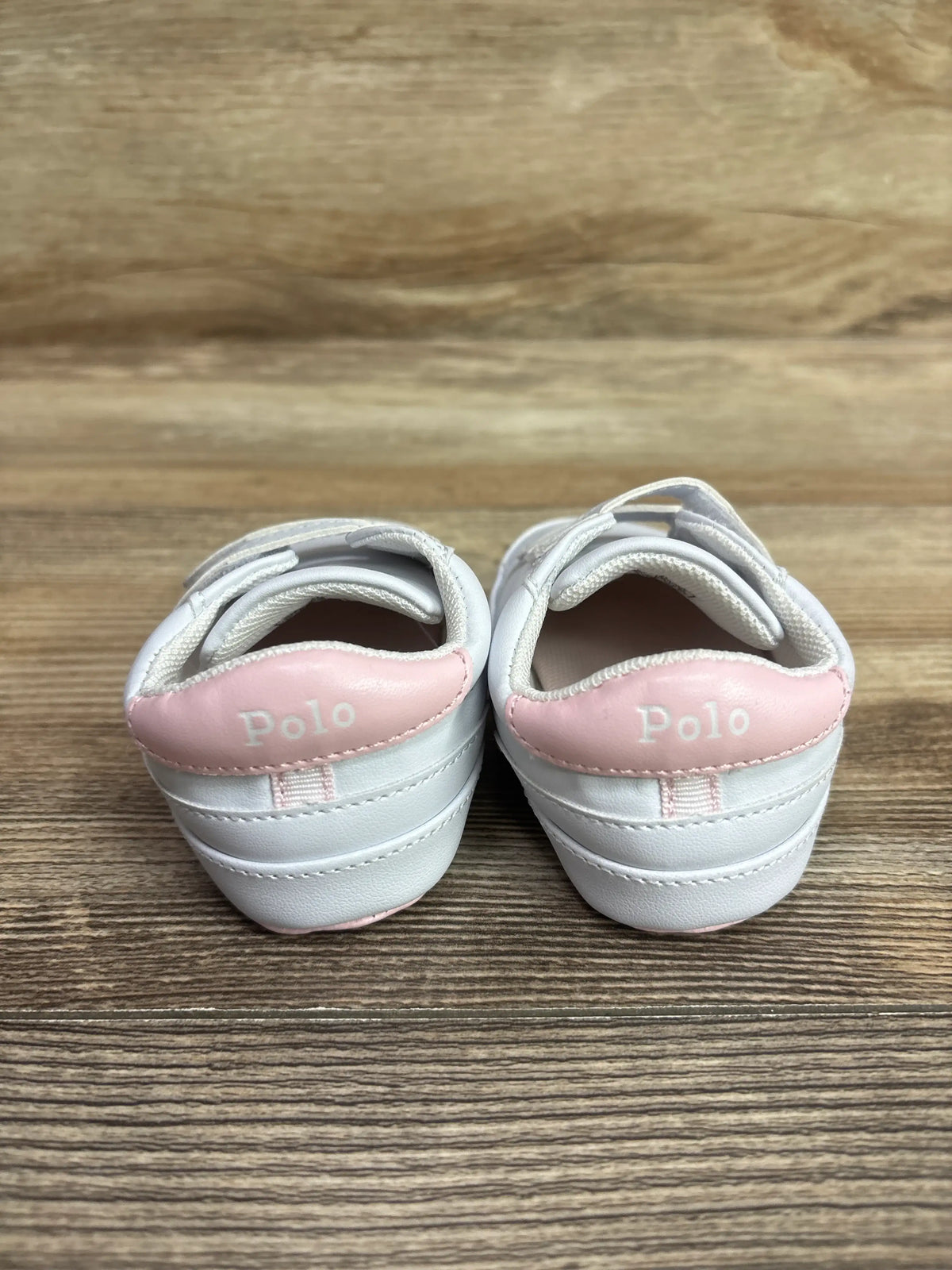 Rear view of Polo Ralph Lauren Heritage Court II EZ crib sneakers in white, showcasing pink Polo heel panels on a wood surface