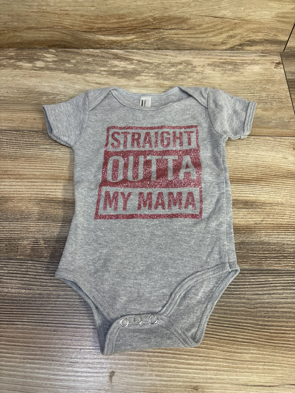 Top-down view of grey American Apparel baby bodysuit showcasing red glitter STRAIGHT OUTTA MY MAMA text with three metal snaps on wood-grain background