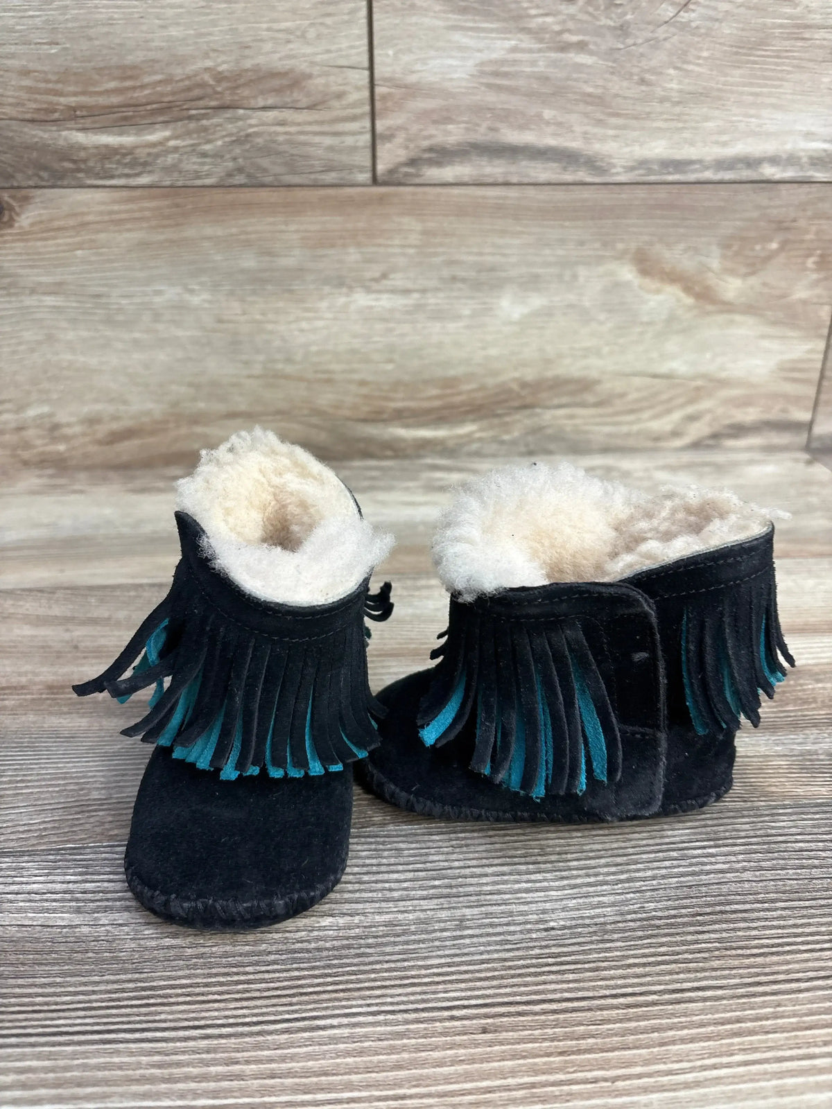 Pair of black toddler moccasin boots with teal fringe and white fleece lining on wood floor