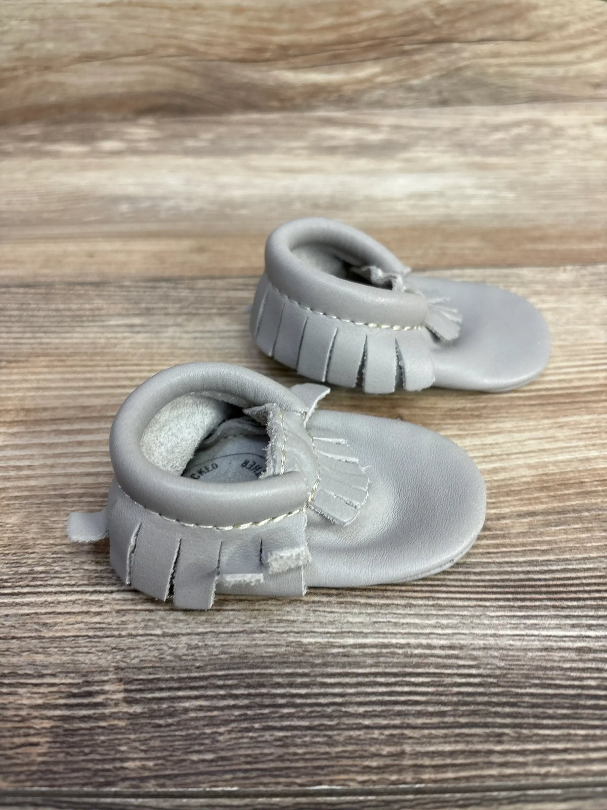 Pair of soft gray baby moccasins with fringe details on a wooden surface
