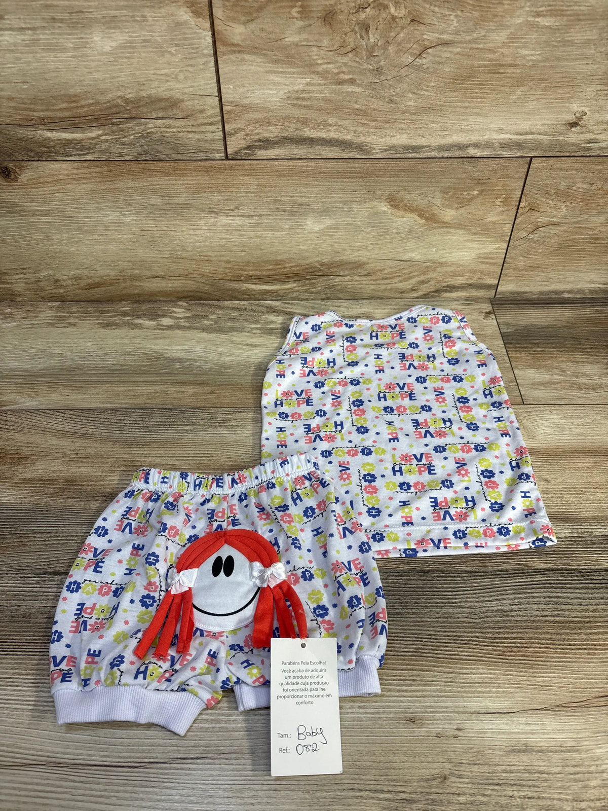 White baby tank top and shorts set with colorful letter-and-flower print and red-haired character applique on wood floor