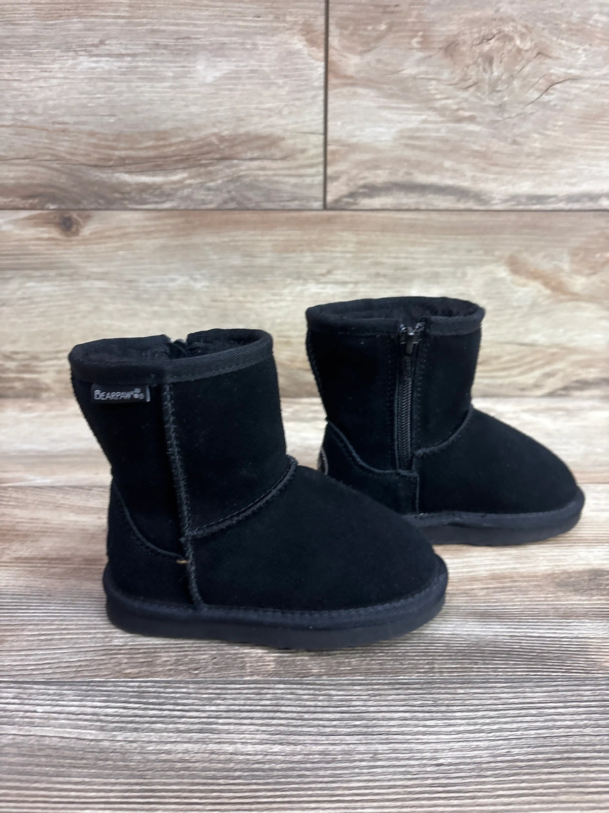Front three-quarter view of BearPaw Eliza boots showcasing side zippers and black uppers on a wood floor
