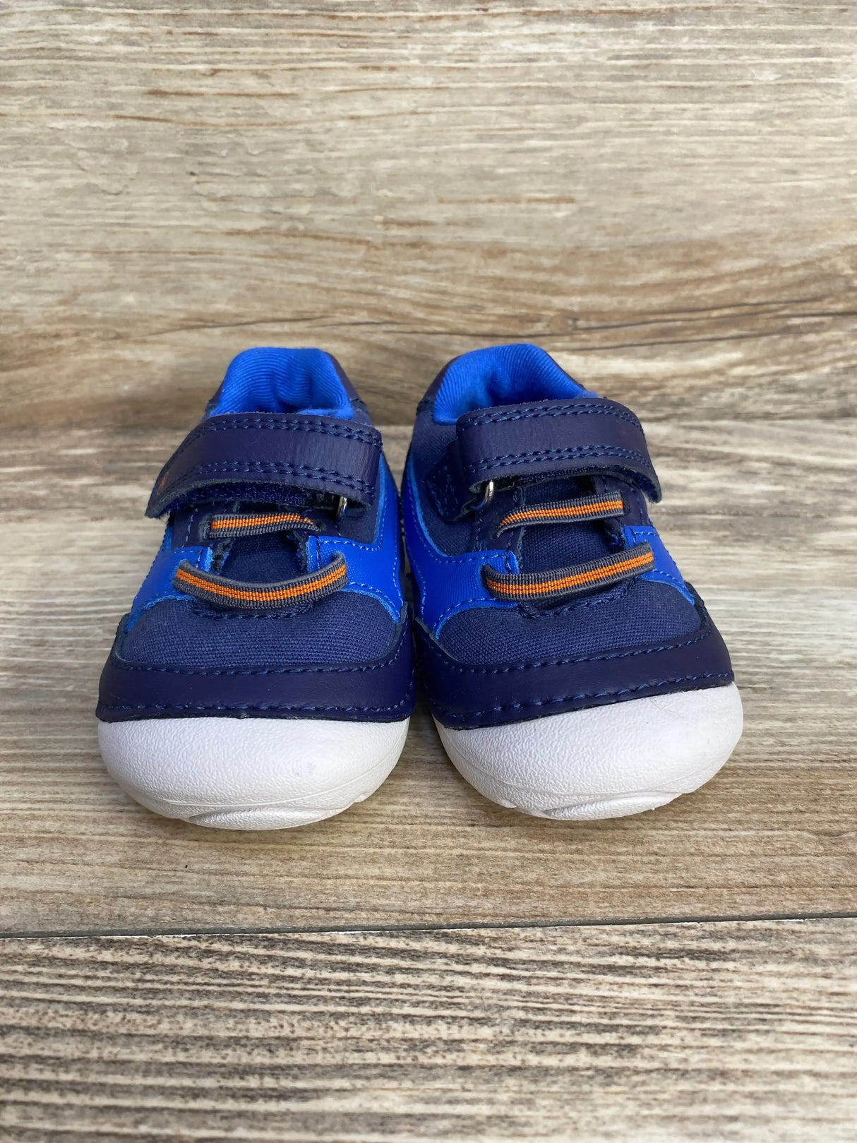 Front view of Stride Rite Soft Motion toddler sneakers in blue showing velcro strap, orange elastic laces and white rounded toe on wood background