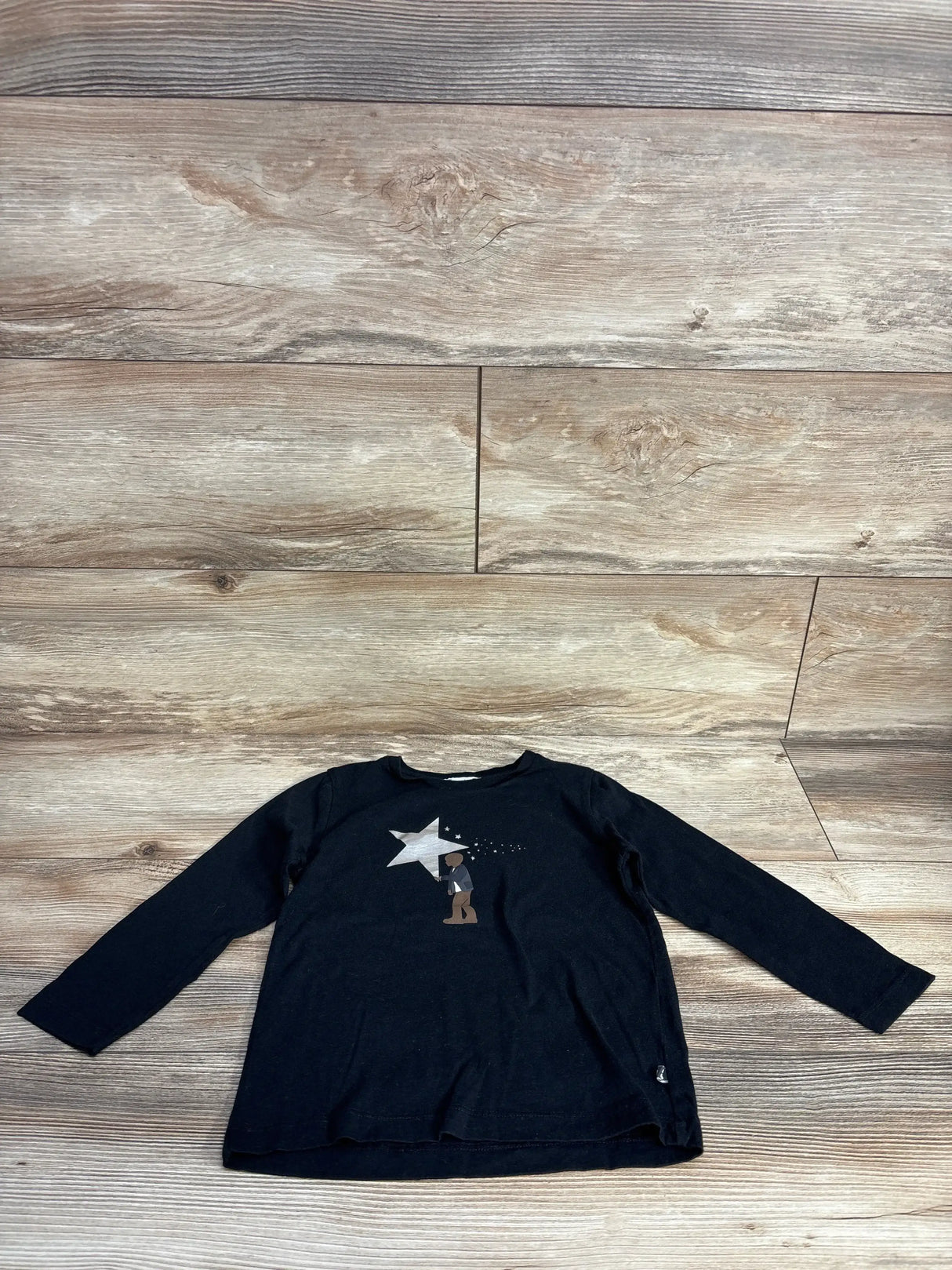 Black long-sleeve child's shirt with graphic of person reaching for large silver star on wood floor