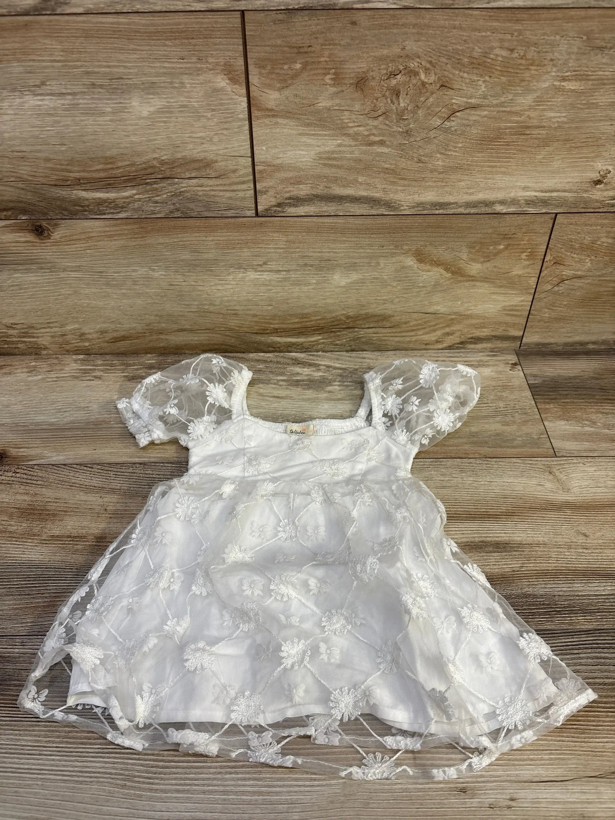 Top down flat lay of Tullabee white embroidered tulle baby dress showcasing puff sleeves and floral lace on wood floor