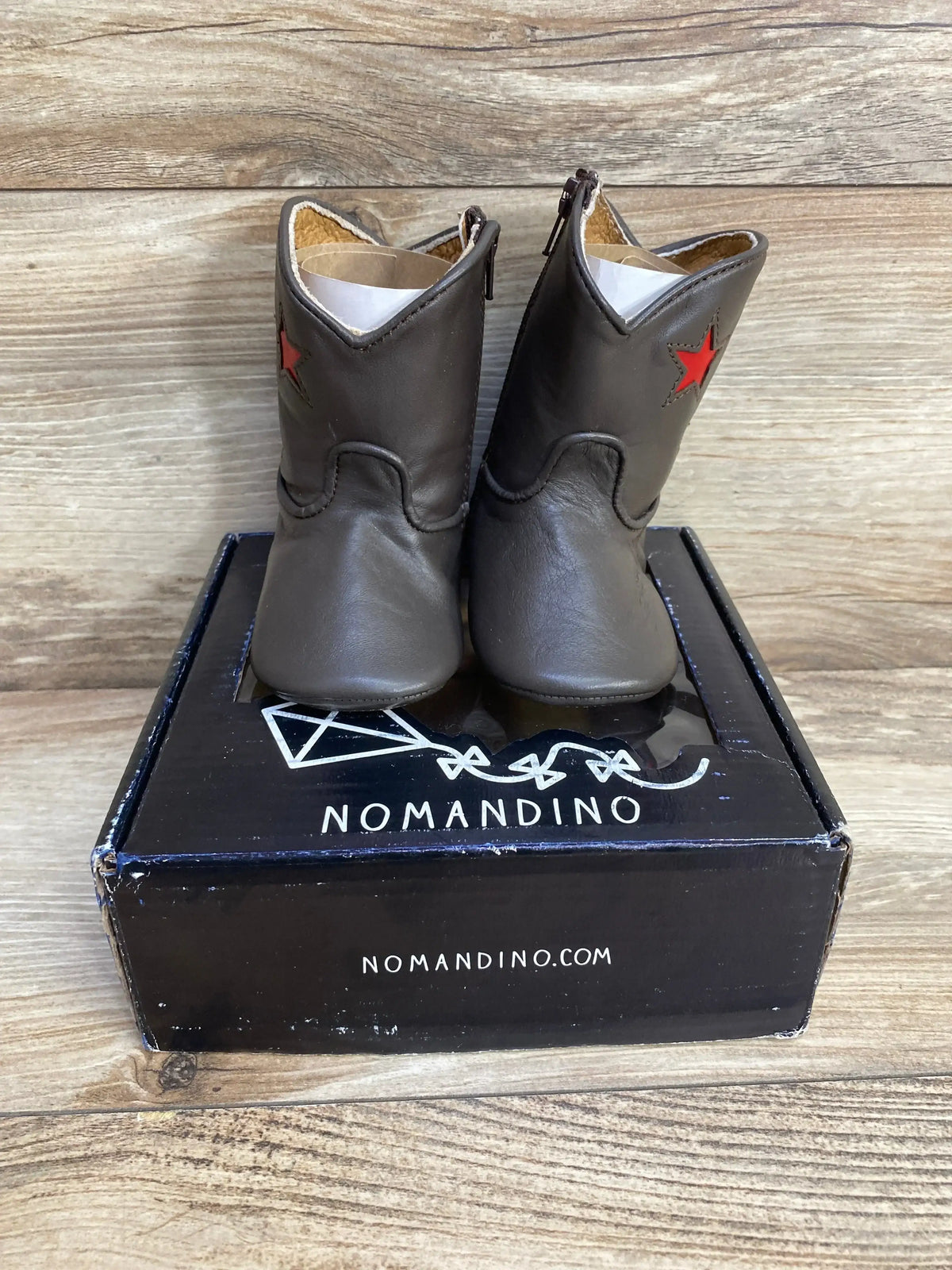 Front view of Nomandino chocolate boots size 2 with red star on shafts on black Nomandino box against wood backdrop