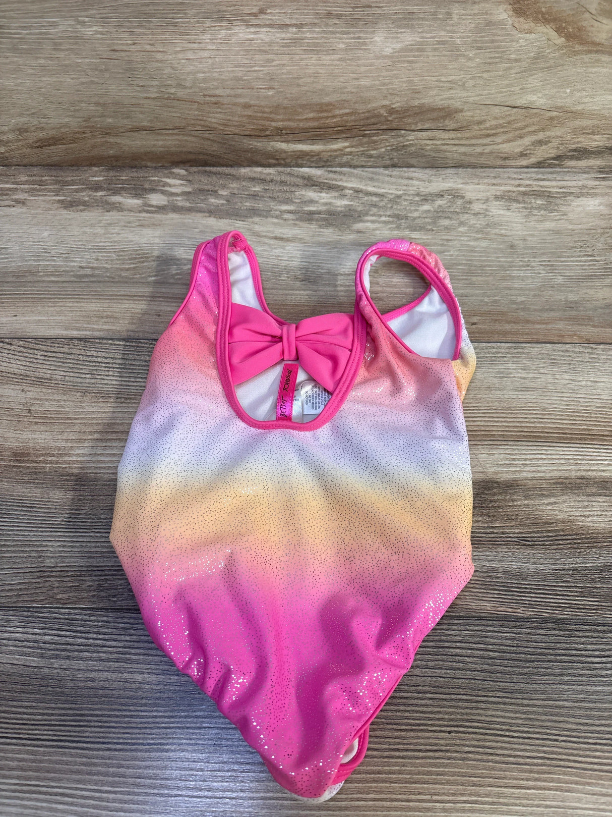 Flat lay of toddler one-piece swimsuit showcasing pink-to-yellow ombre, glitter dot finish, and back bow detail on wood background