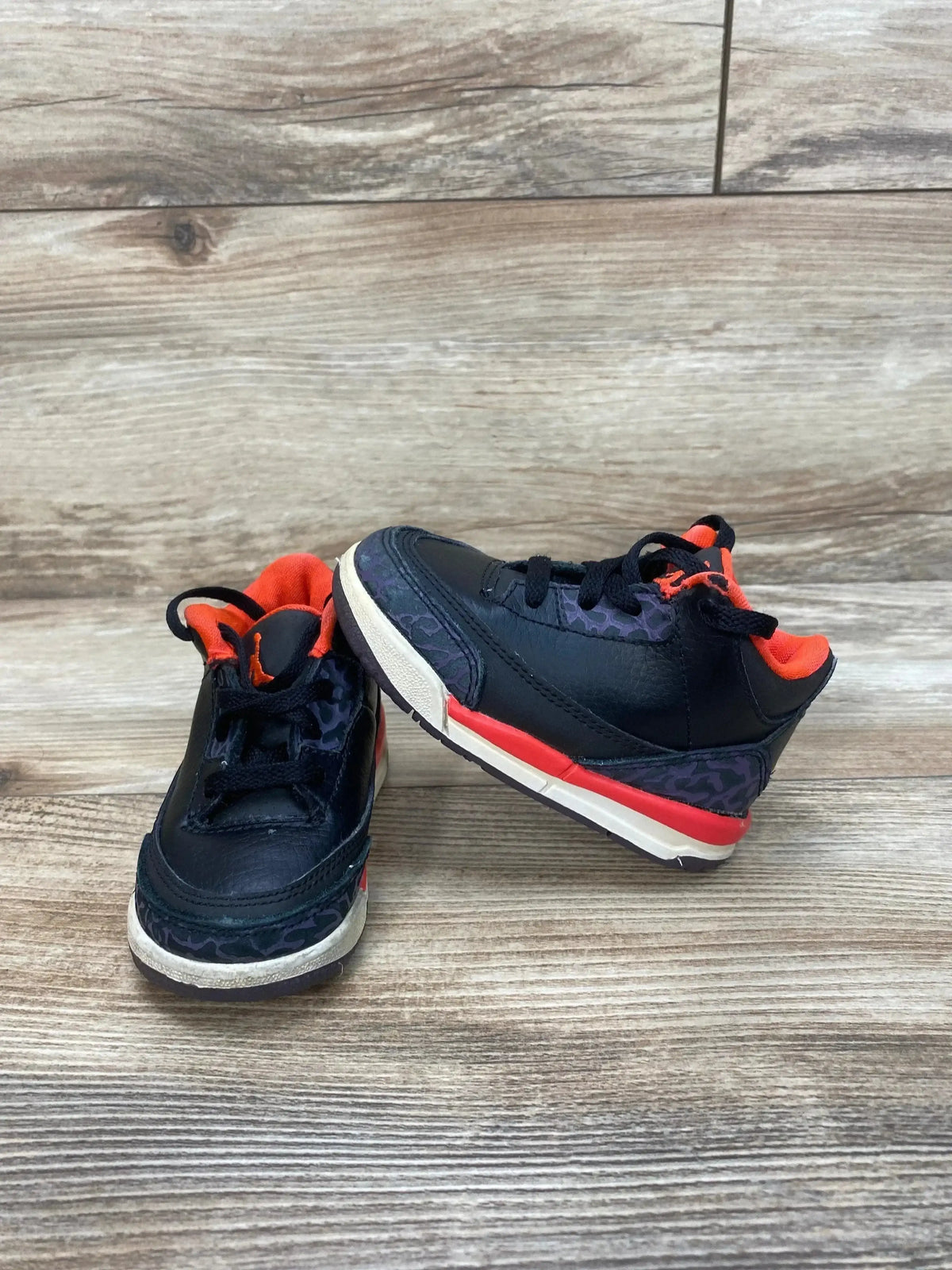 Front three-quarter view of toddler Air Jordan 3 Retro, black with crimson lining, elephant-print accents on wood floor