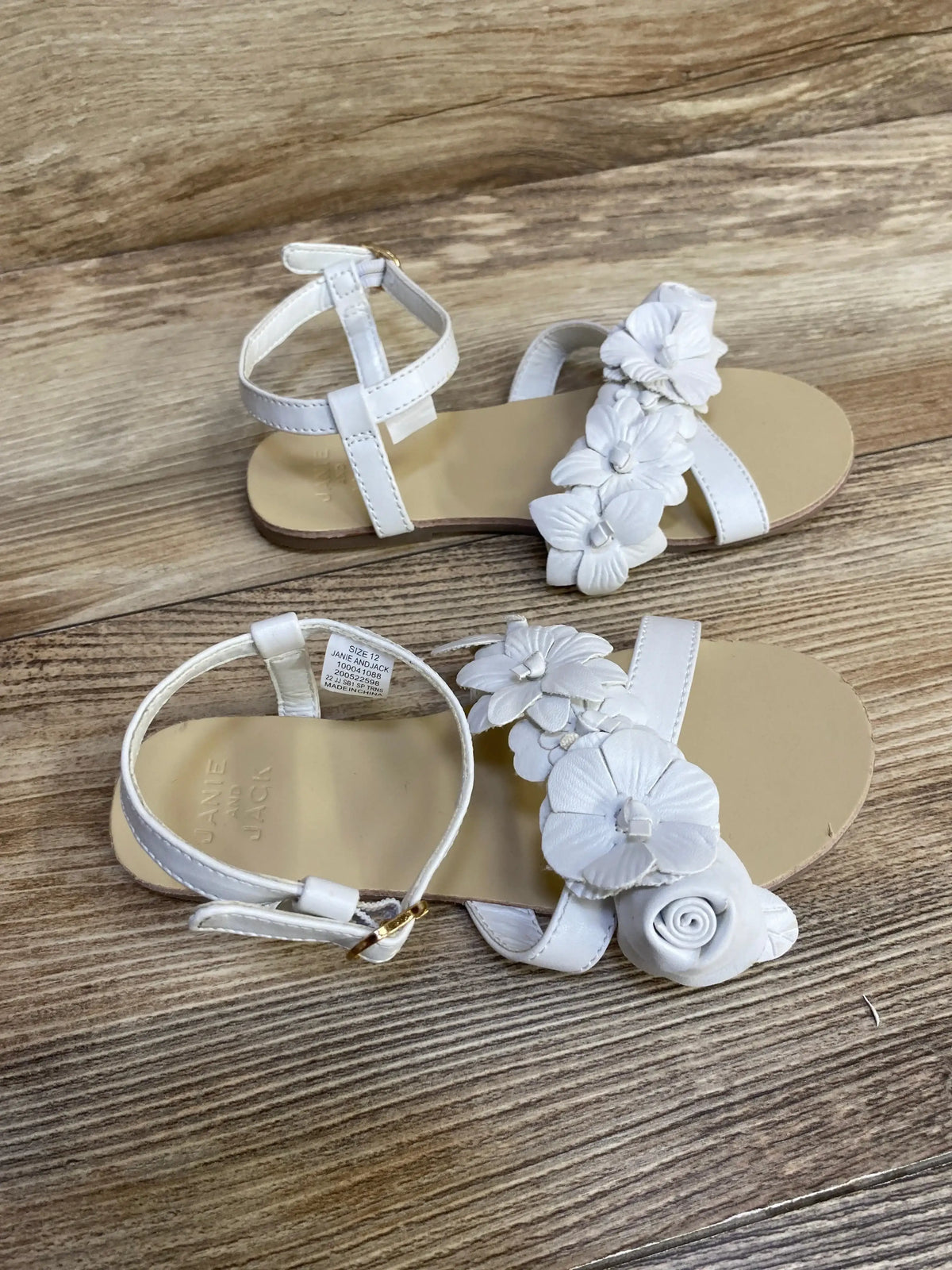 Top-down view of Janie and Jack white sandals with flower applique strap, ankle buckle, size 12 tag on wood floor