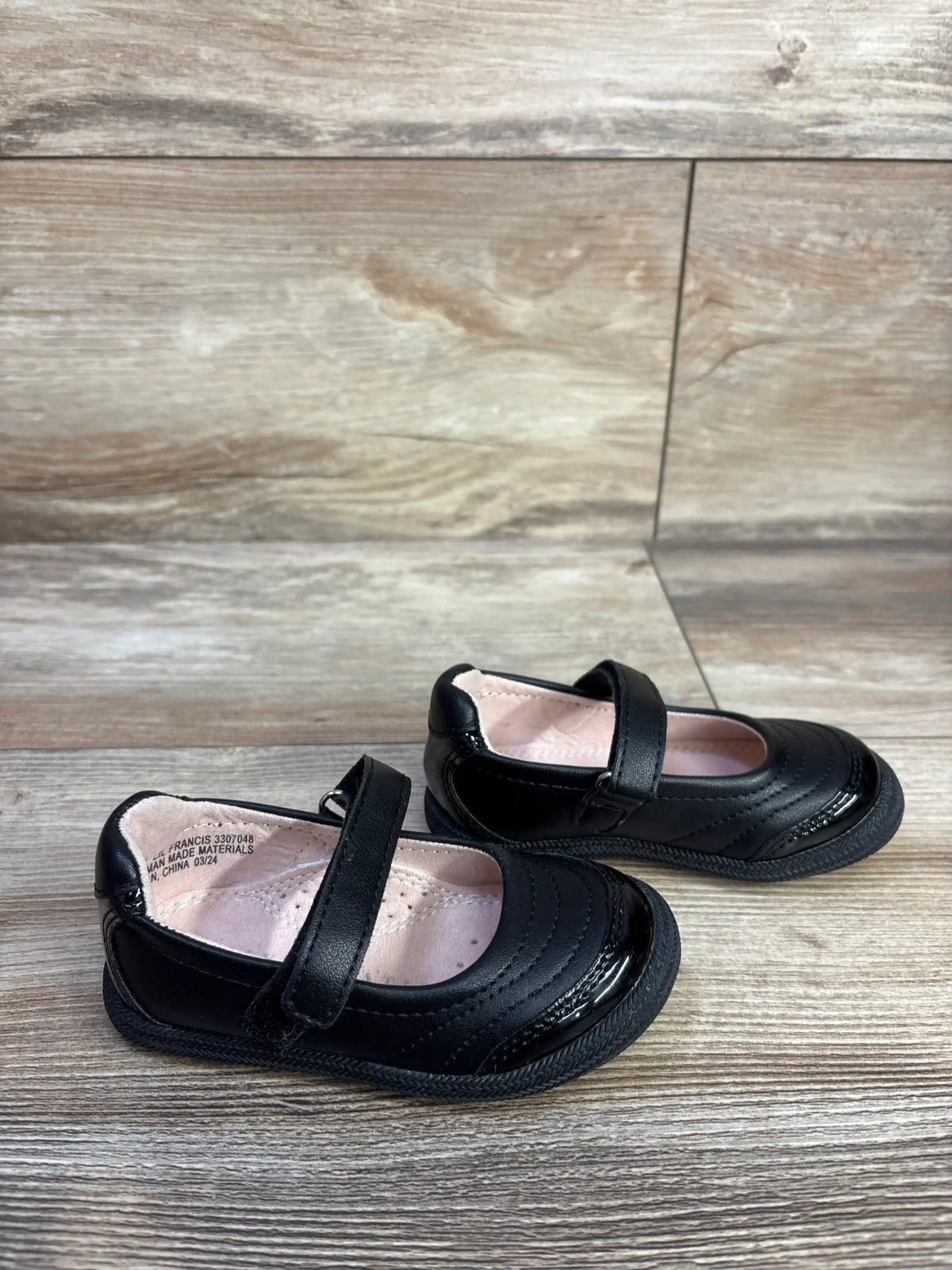 Angled view of Kelly and Kate Mary Jane toddler flats in black showing strap closure and glossy toe on light wood floor