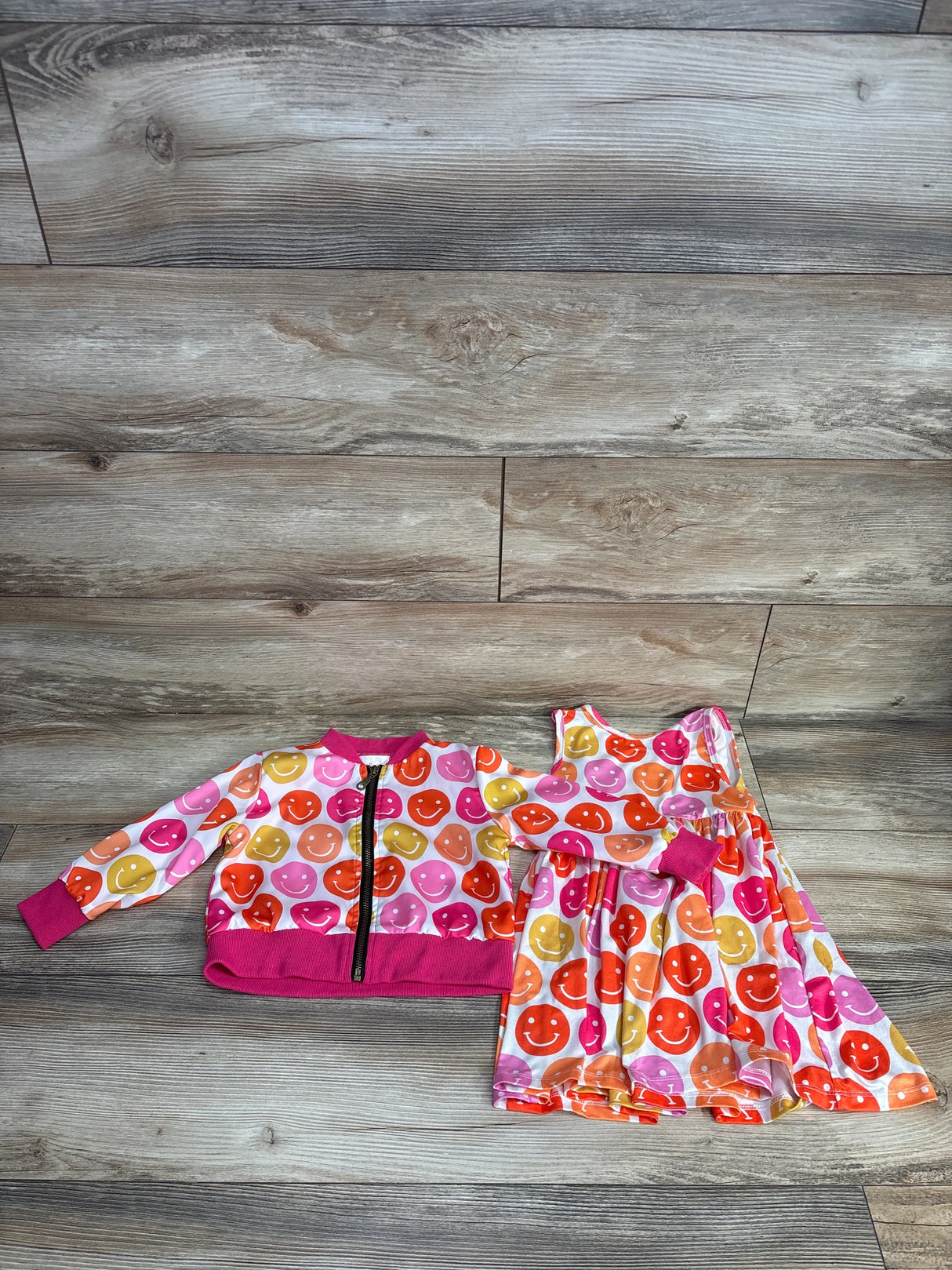 Kids clothing set with colorful smiley face print including a pink-trimmed zip jacket and matching sleeveless dress on wooden floor