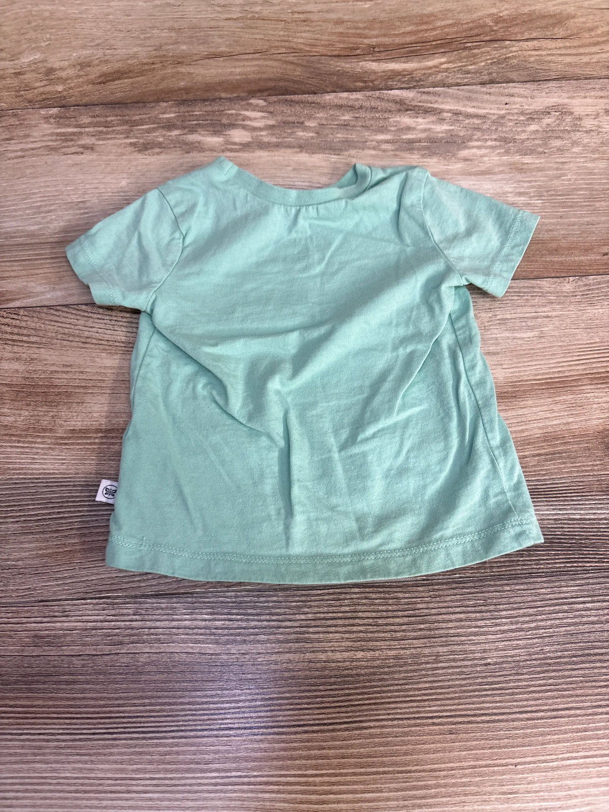 Front view of Honest Baby solid green shirt, size 3-6m, showcasing short sleeves and crew neckline on a wood background