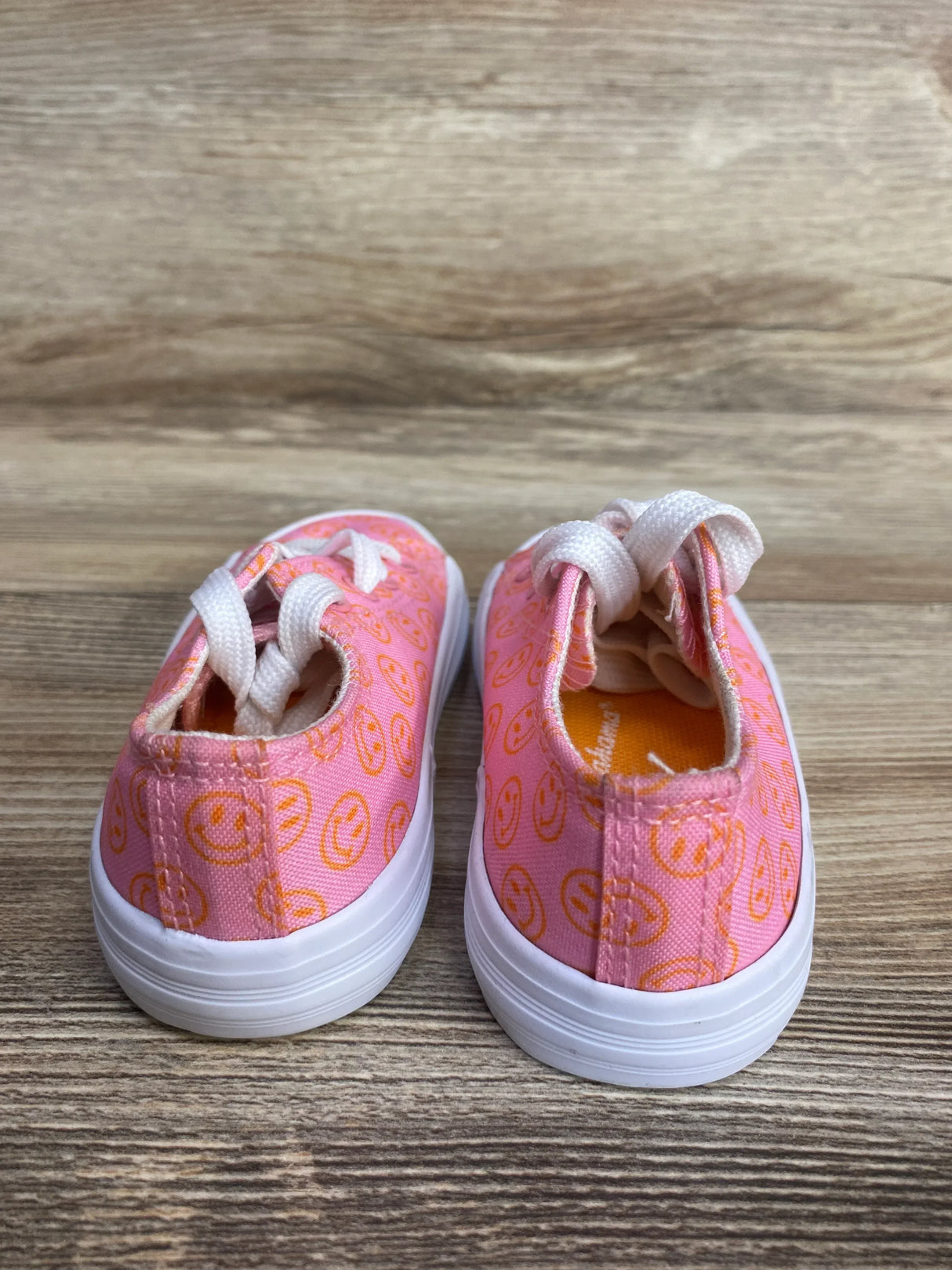 Pink toddler sneakers with orange smiley face patterns and white laces on wooden floor