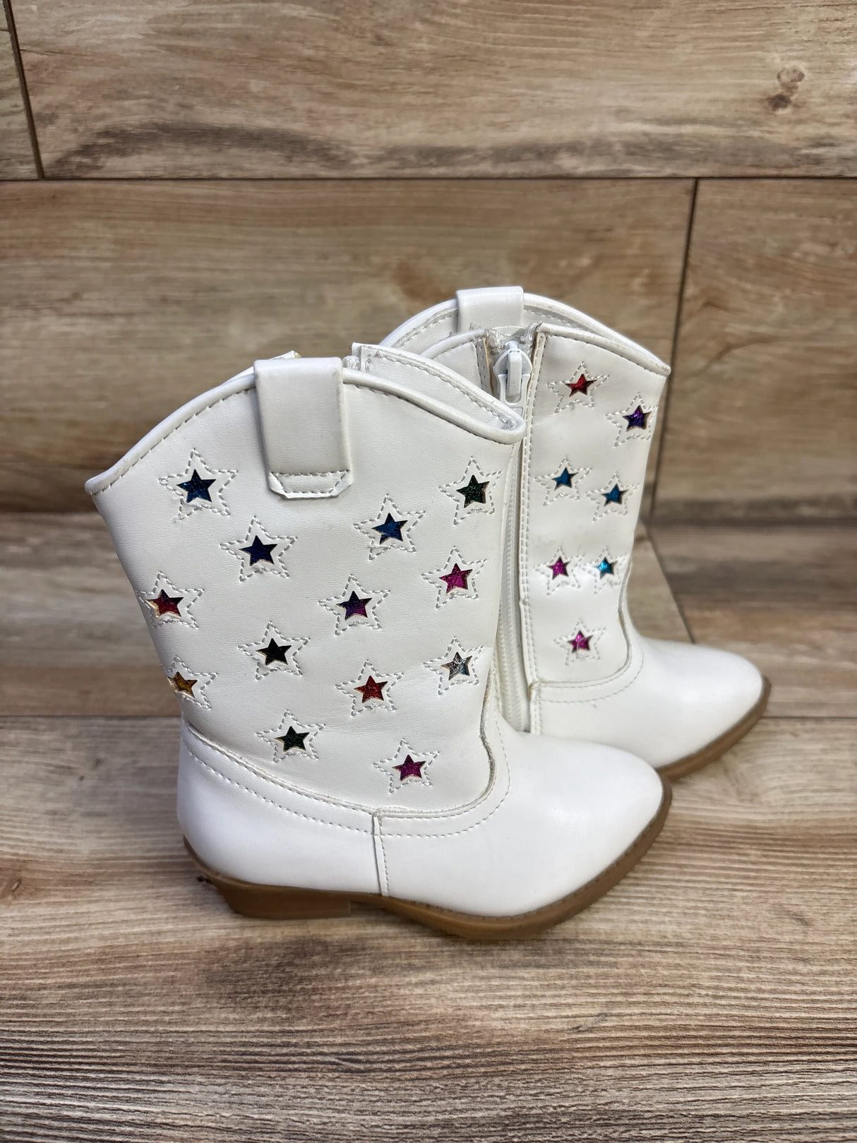 Side view of toddler Tucker and Tate white cowgirl boots showing multicolored star insets and side zipper on wood floor