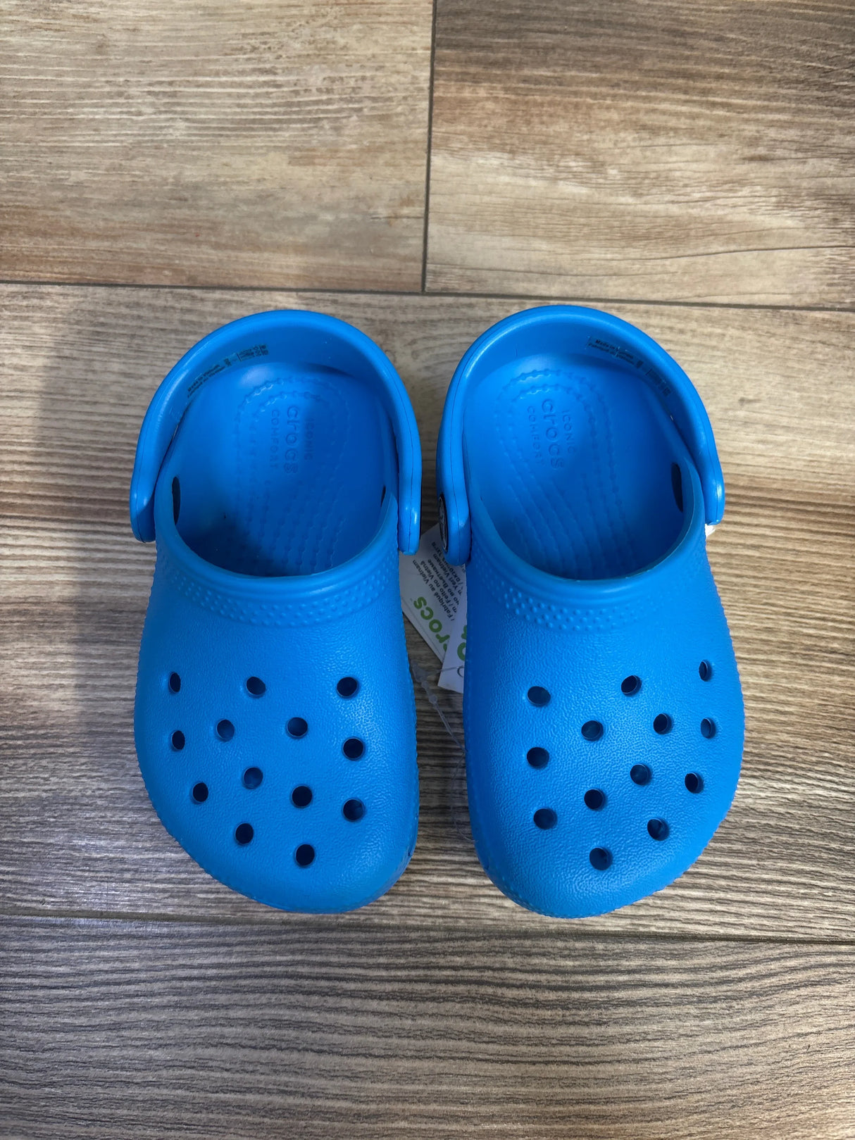 Top-down view of toddler blue Crocs Classic clogs showcasing toe ventilation holes and heel straps with wood floor background