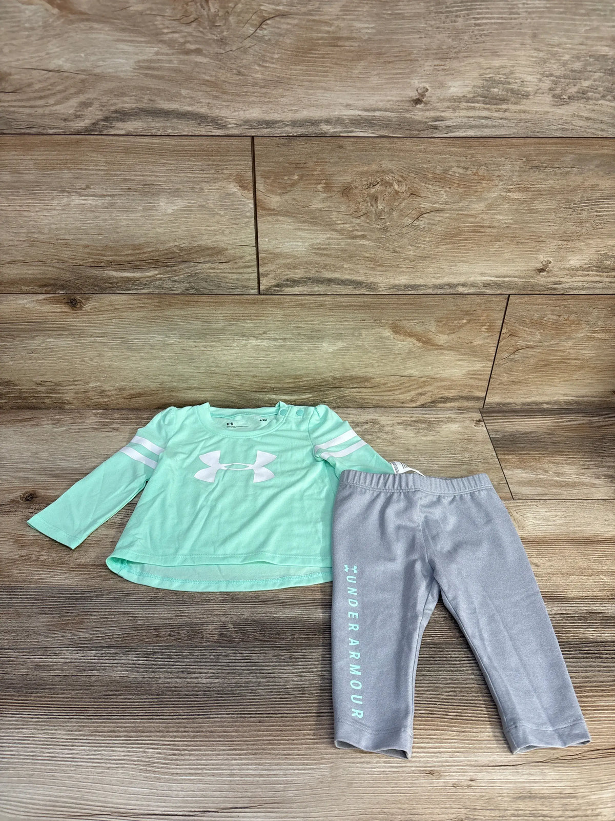 Top view of Under Armour two-piece long sleeve shirt and pants set showcasing teal shirt and gray pants with logos on wood background