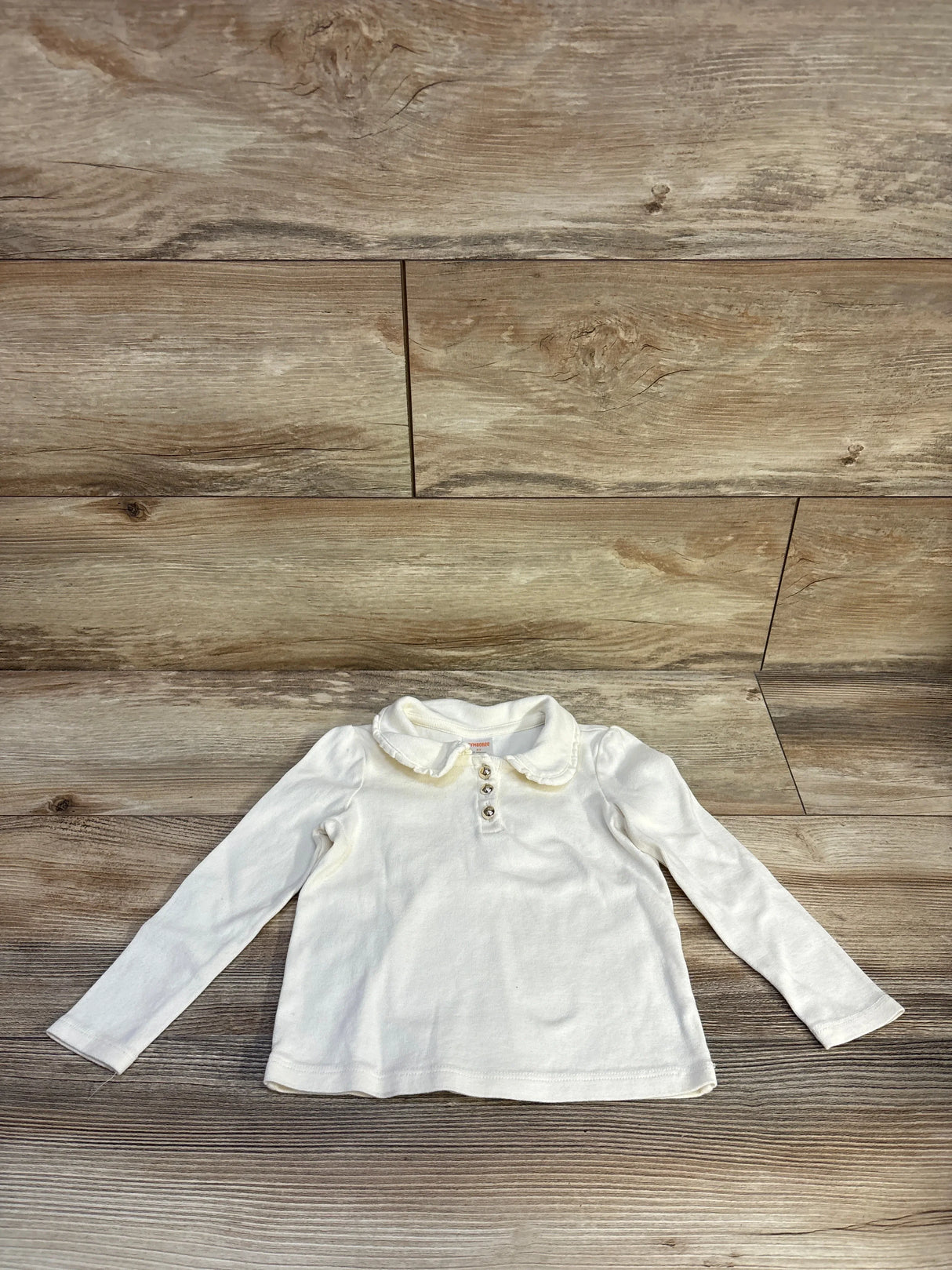Flat lay of cream Gymboree long-sleeve Peter Pan collar top with ruffle collar and three buttons on wood floor, size 3T