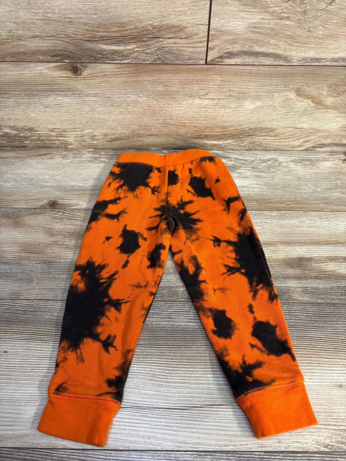 Front view of orange Halloween joggers with black tie-dye pattern and cuffed ankles on a wood-look floor