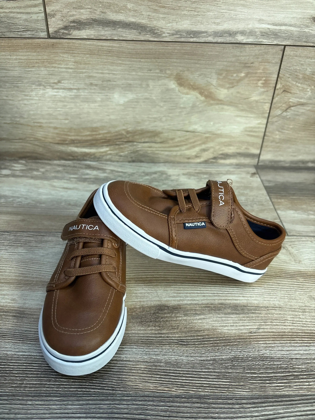 Angled view of Nautica Calloway faux leather brown sneakers in size 10C with lace-up fronts and white soles on wood-look floor