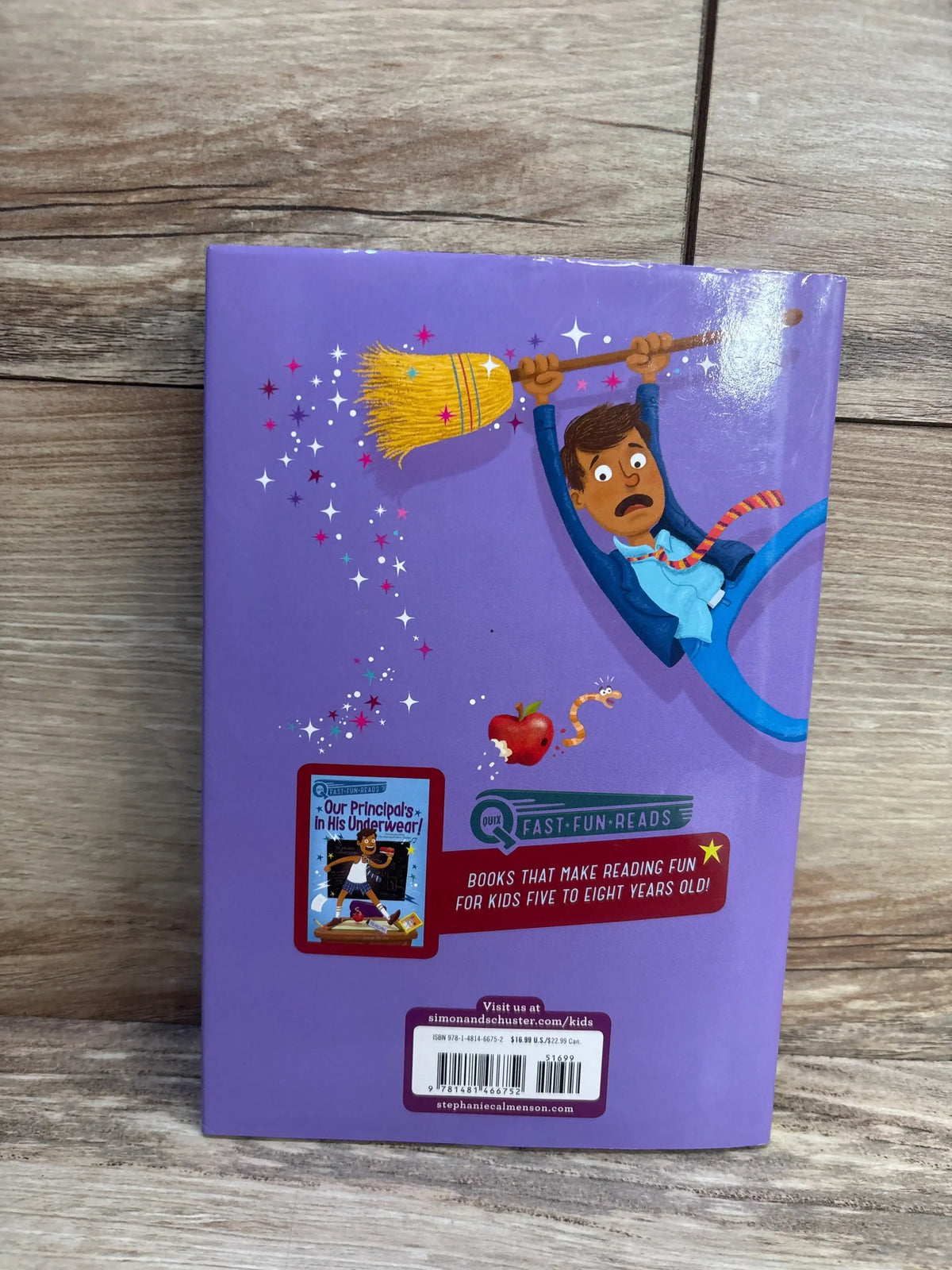 Back cover of Our Principal Breaks a Spell! hardcover showing a cartoon principal hanging from a broom with sparkles, publisher blurb ‘Books that make reading fun for kids five to eight years old’ on a wood floor background