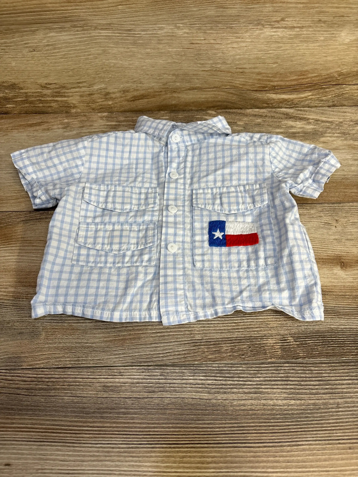 Front view of blue gingham baby button-up shirt showcasing two chest pockets and embroidered Texas flag patch on wood surface