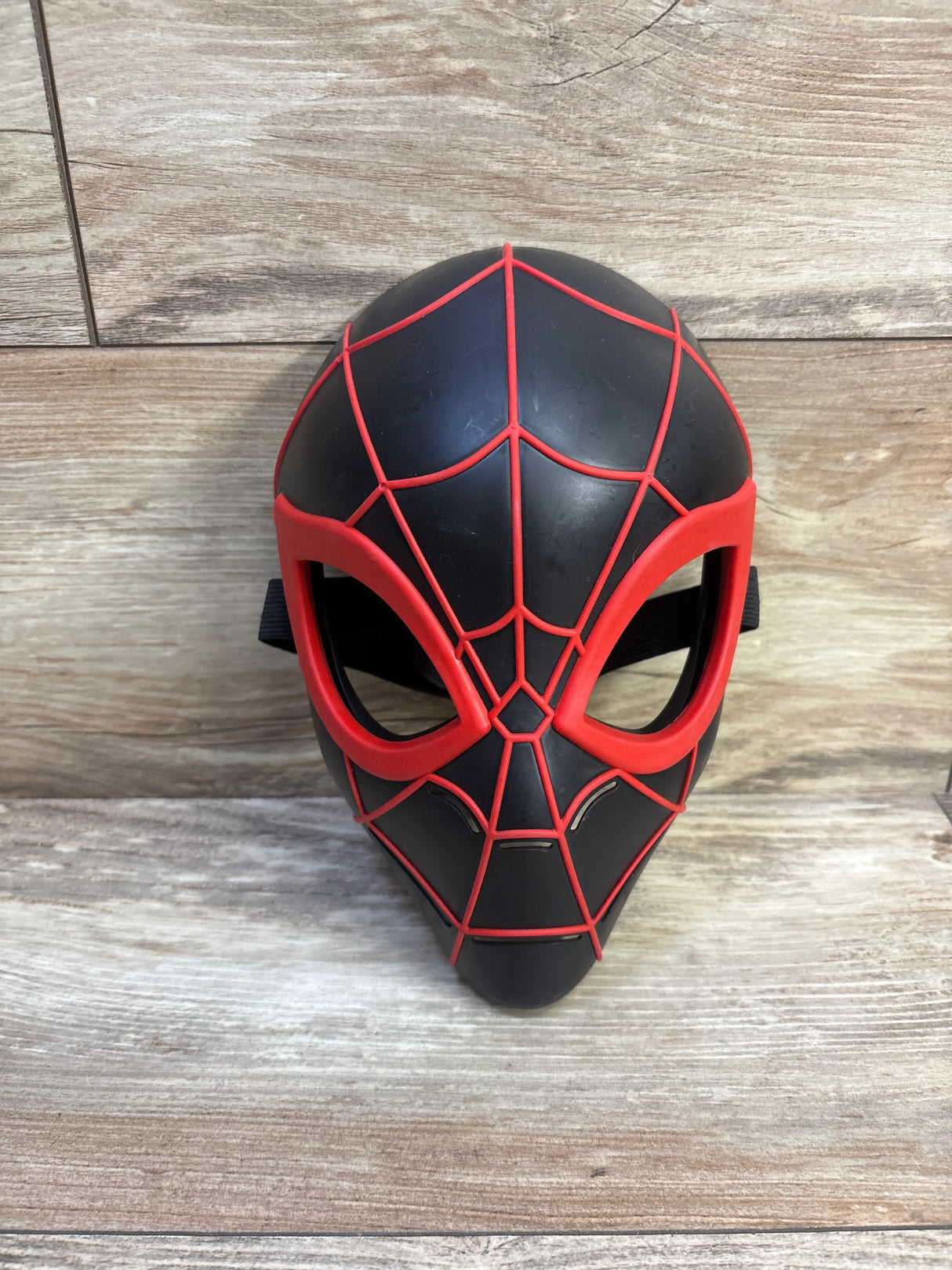 Front view of Miles Morales Spider-Man mask showing black molded face with red webbing and elastic strap on wood background