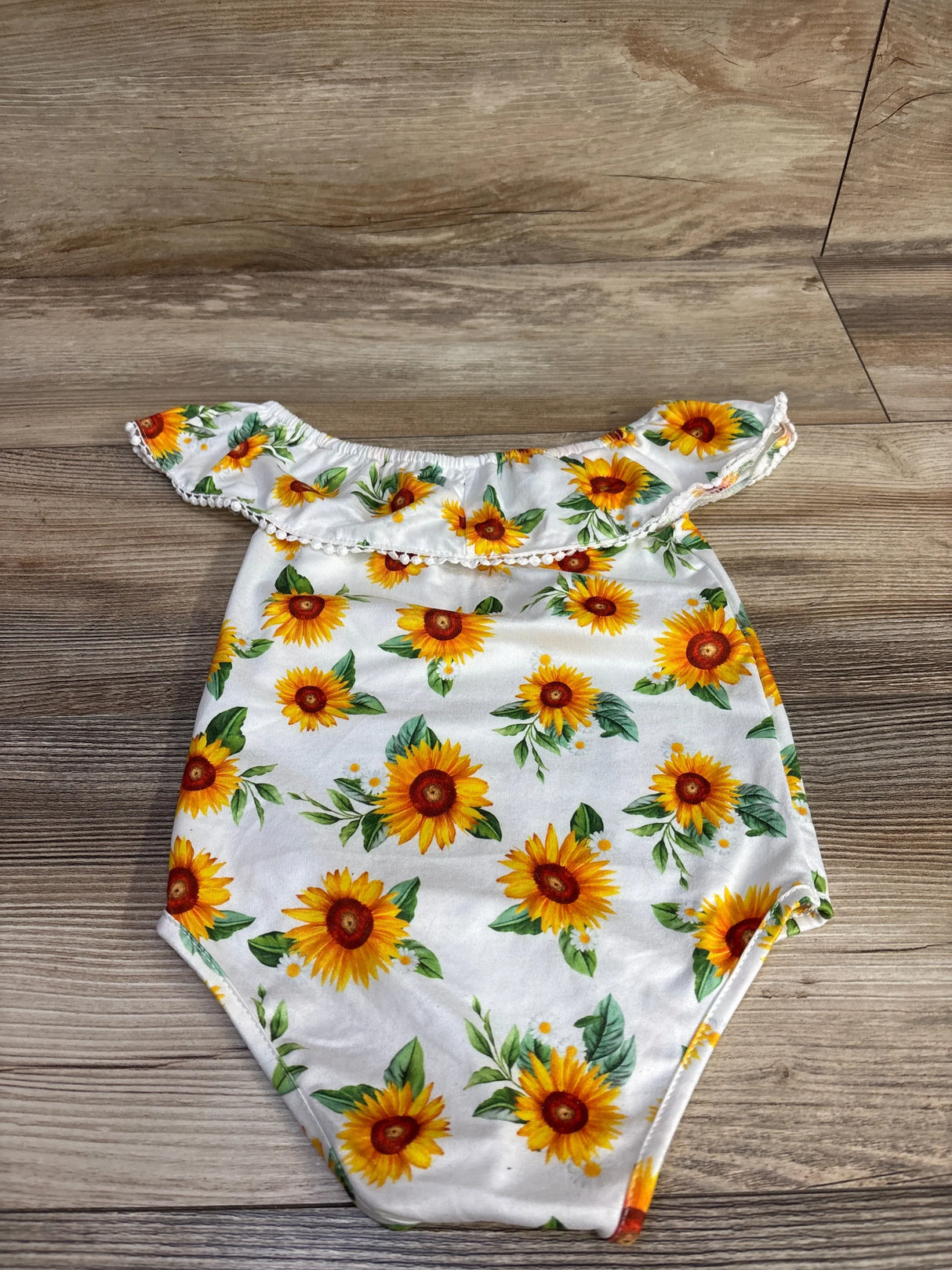 Flat lay of white off-shoulder baby romper showcasing sunflower print and ruffle trim, size 12 months, on wood floor