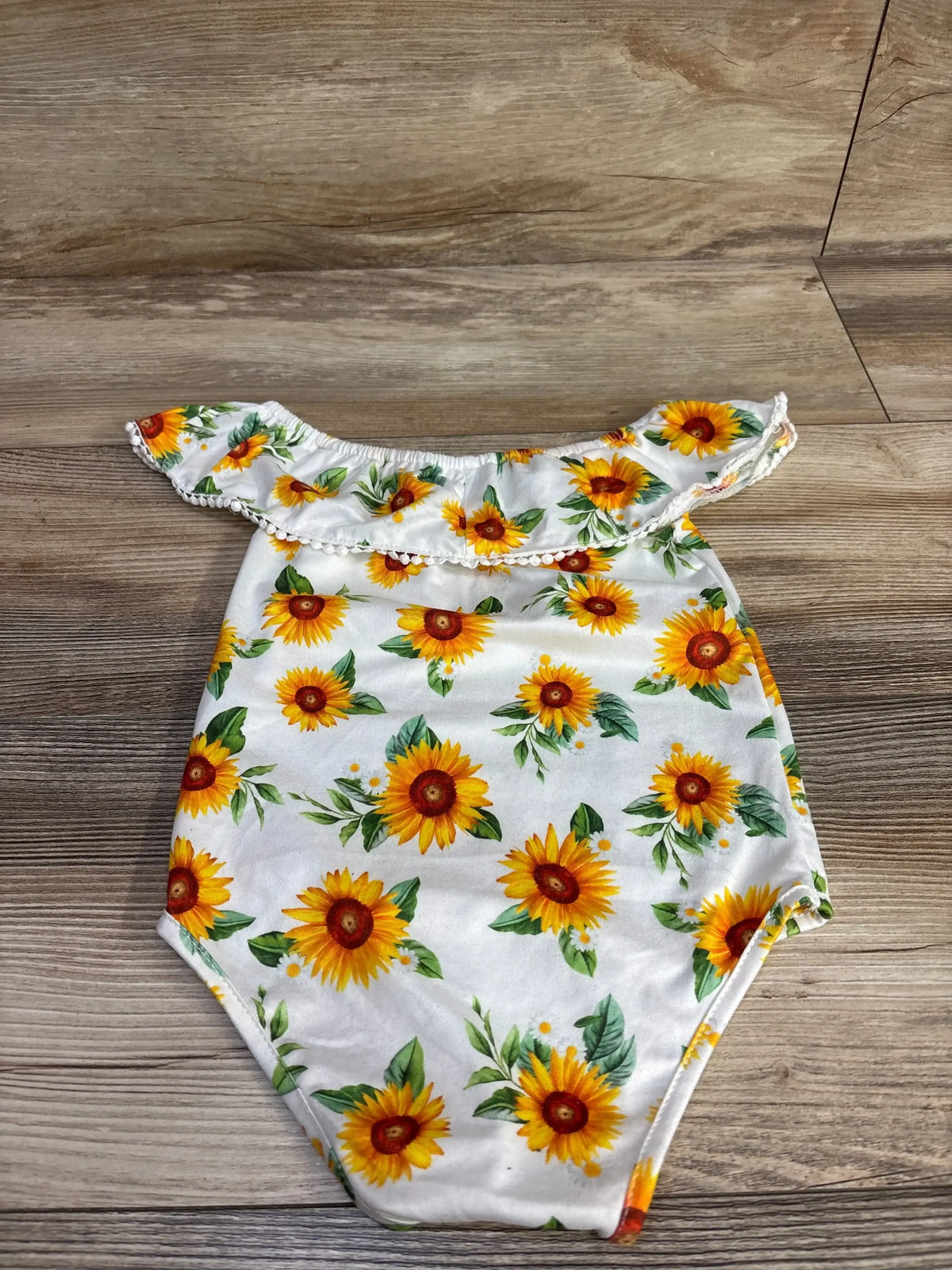 Flat lay of white off-shoulder baby romper showcasing sunflower print and ruffle trim, size 12 months, on wood floor