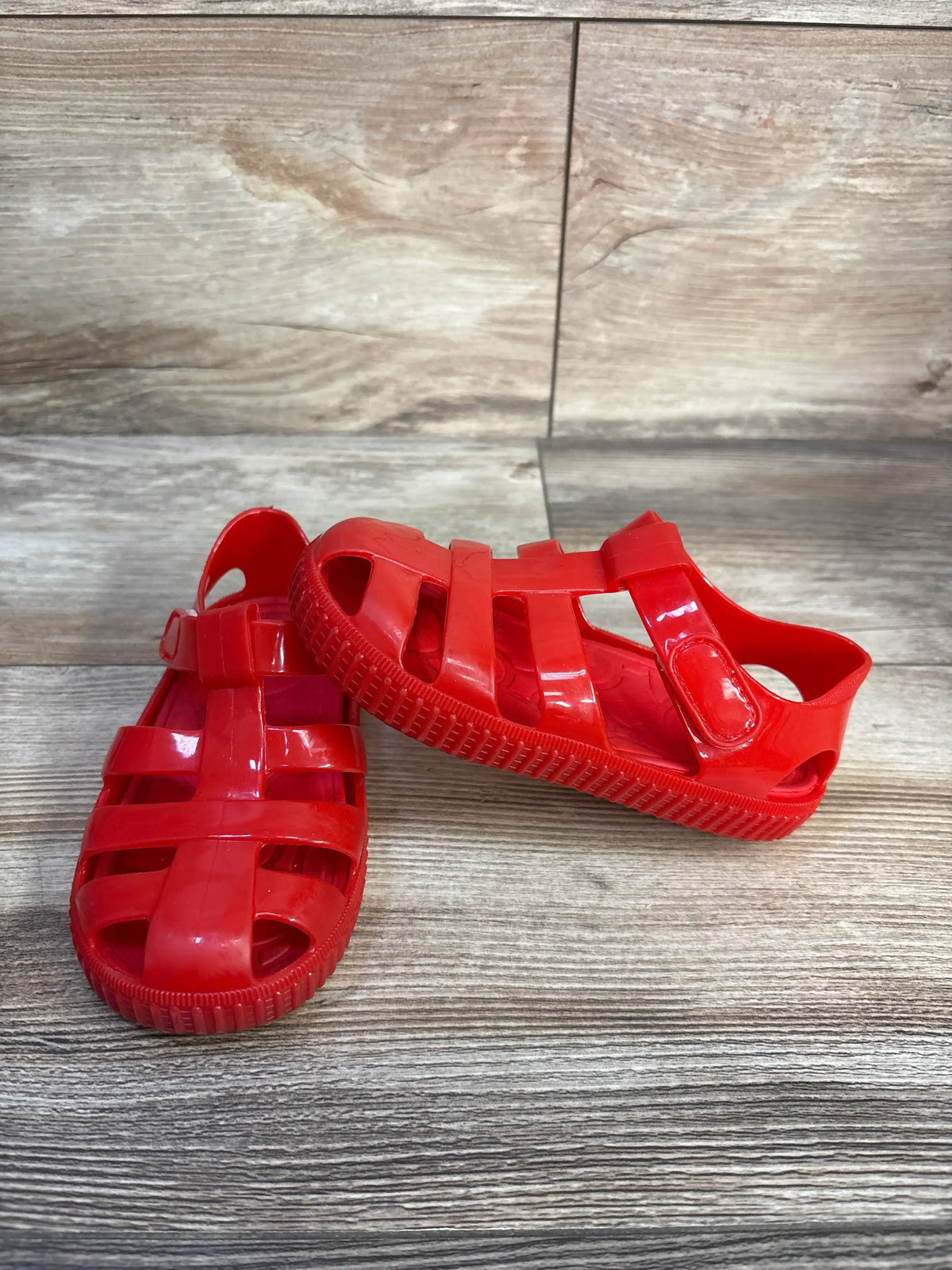 Angled view of red jelly fisherman sandals, size 8/9C, showcasing hook and loop closure on a wood-look background