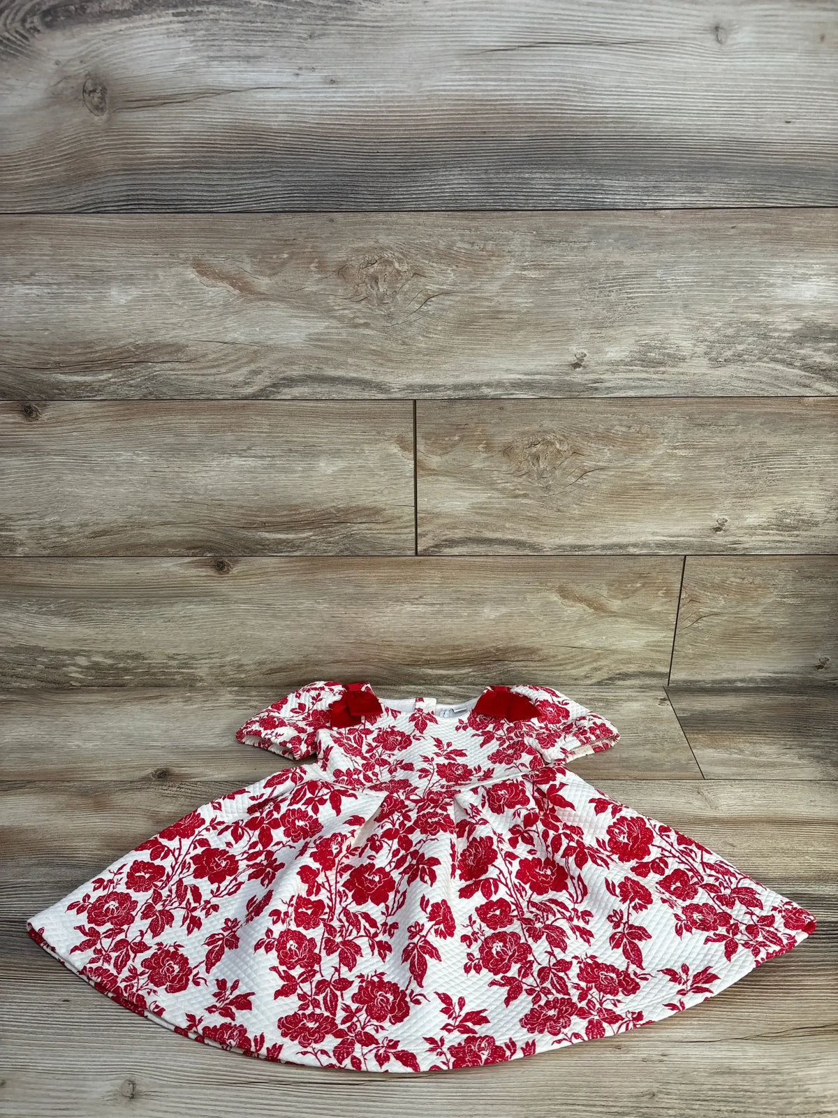 Flat lay of Janie & Jack toddler quilted Maroon Berry floral dress showing red rose print, velvet bow accents and puff sleeves on wood floor background