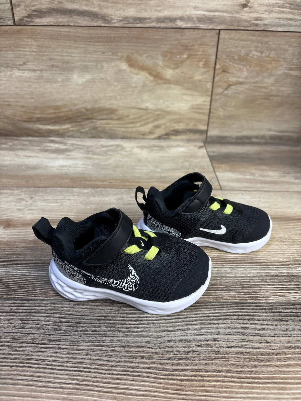Angled side view of Nike Revolution 6 toddler sneaker with black mesh, white patterned Swoosh and neon laces on wood floor