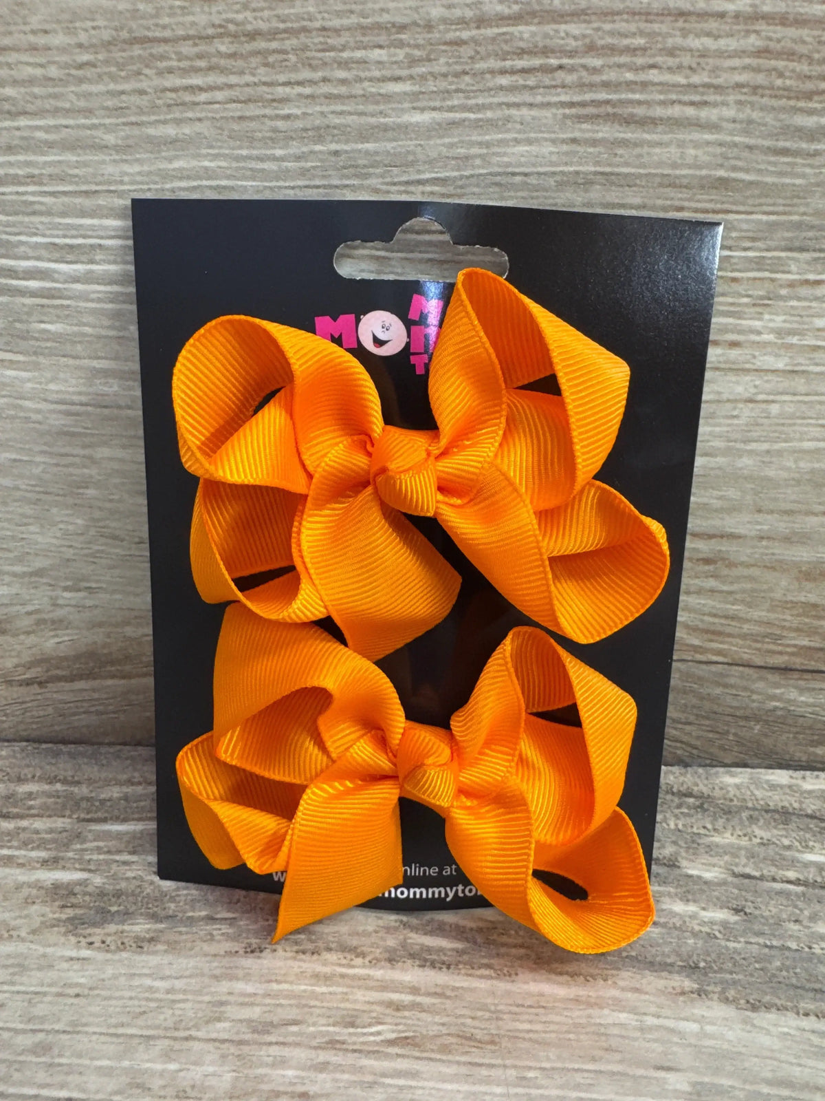 Front view of two medium orange grosgrain hair bows on a black display card against light wood background