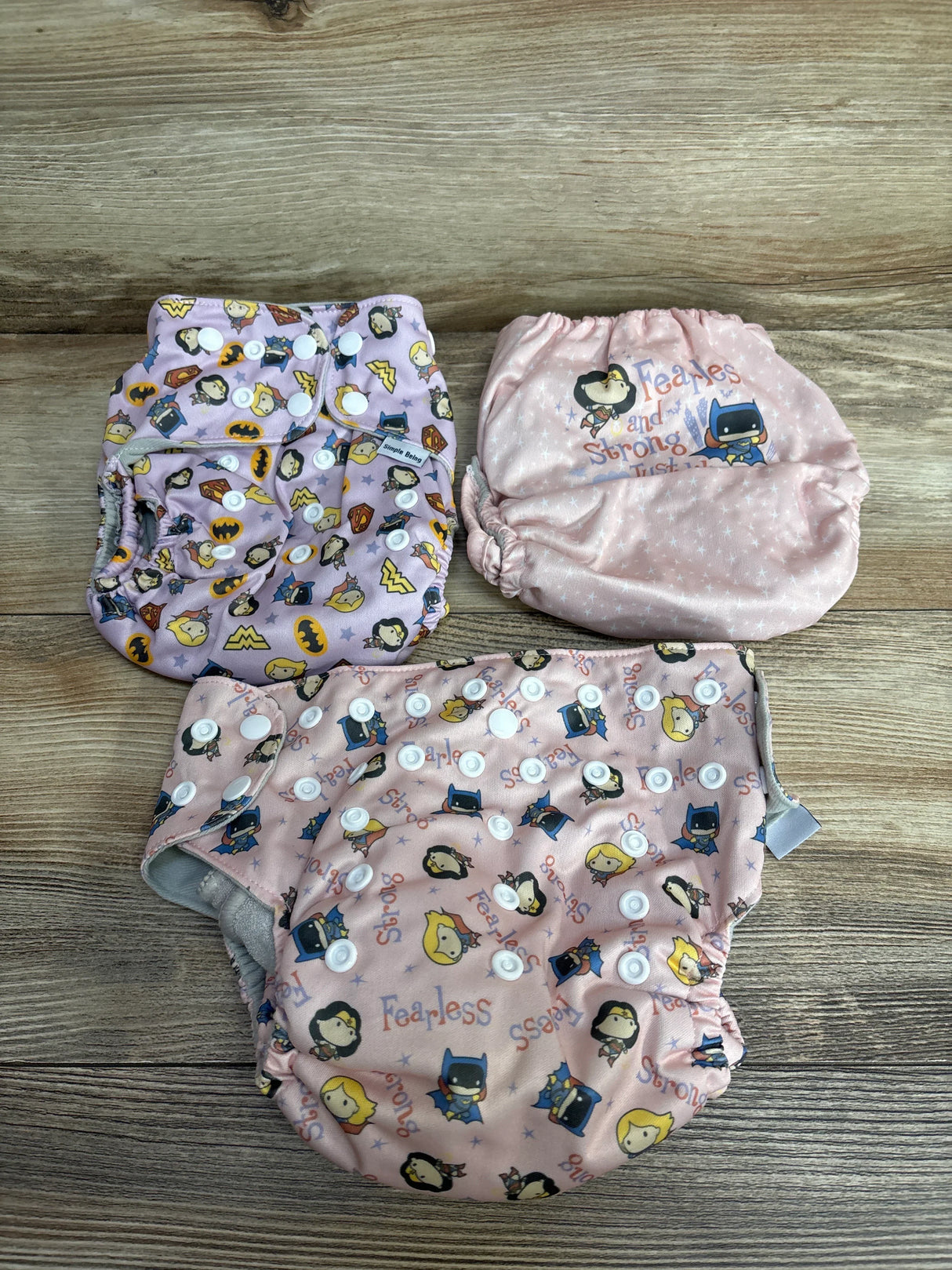 Three reusable cloth diapers with superhero patterns and snap closures on a wood background