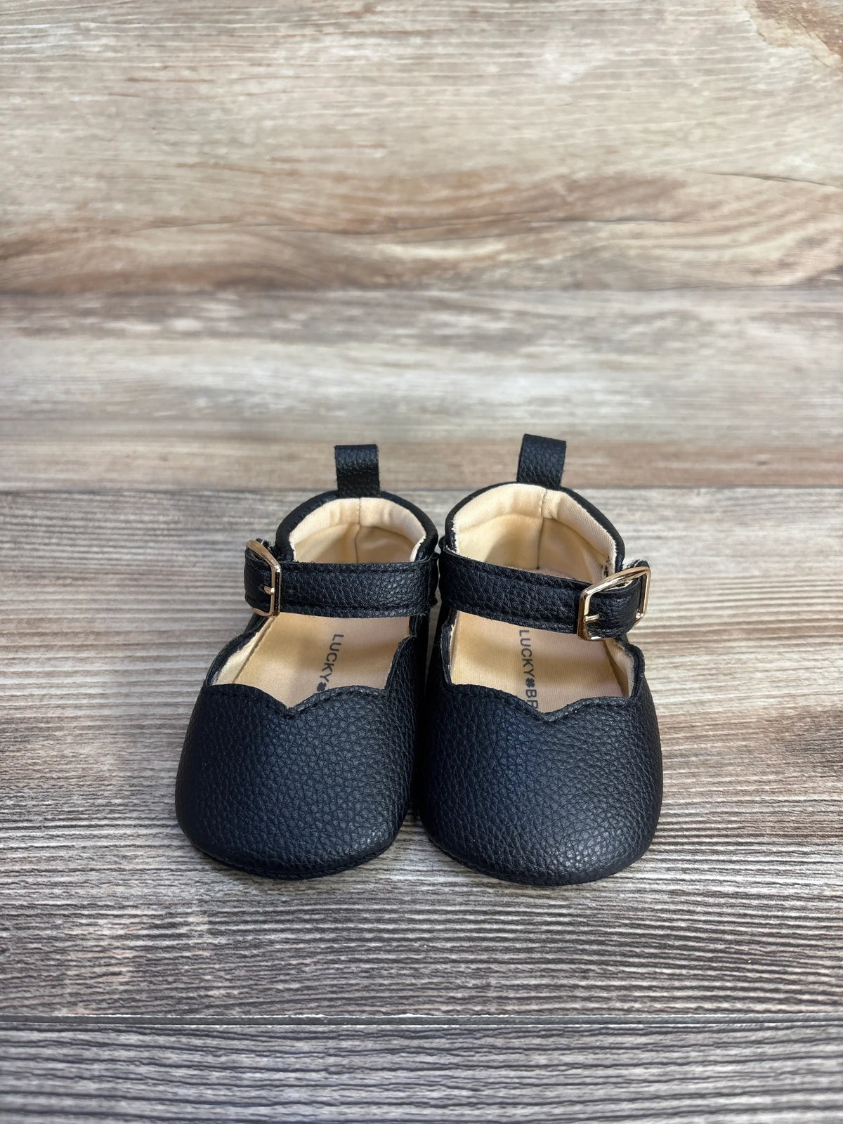 Lucky Brand black leather Mary Jane crib shoes for babies on a wooden surface.