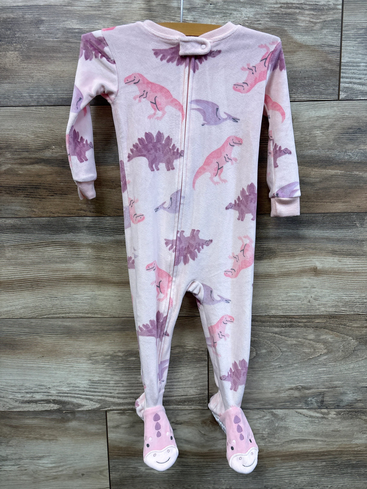 Front view of Carter’s pink dinosaur footed blanket sleeper size 18M showing all-over pink and purple dinosaur print and smiling dinosaur footies on wood background
