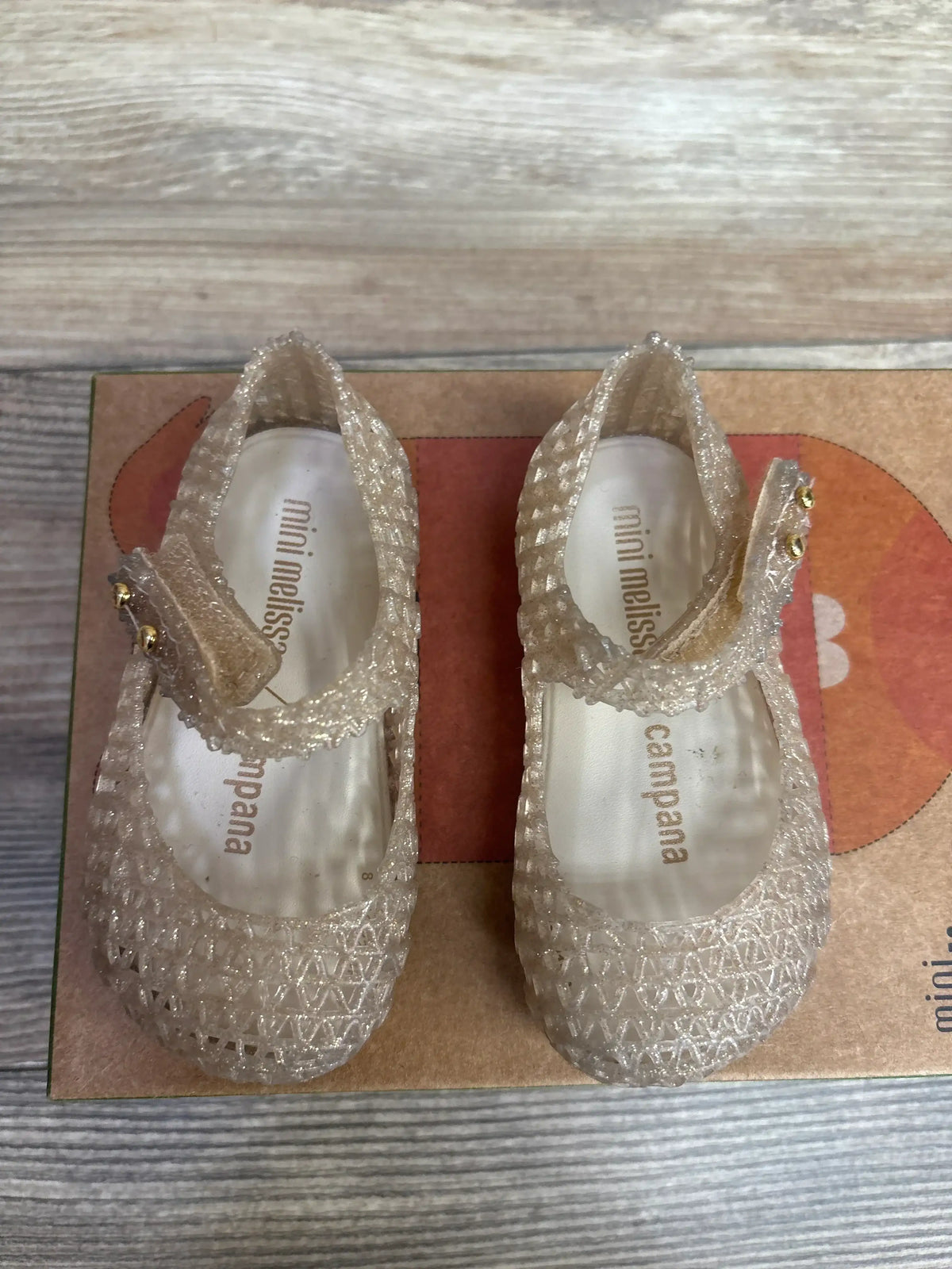 Top view of Mini Melissa Campana glittery clear jelly Mary Jane shoes with woven texture on original orange box on wood plank floor