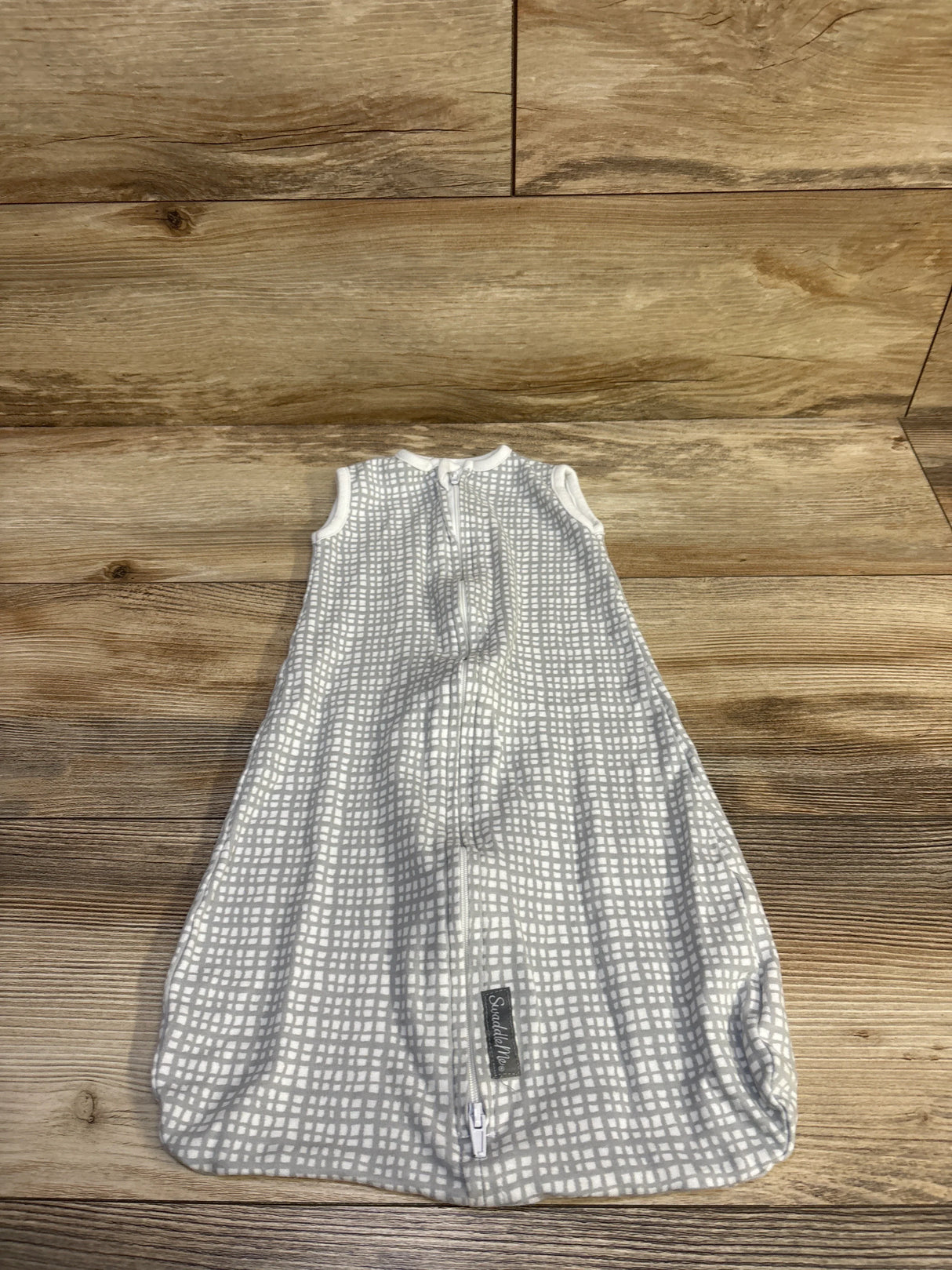 Flat lay of SwaddleMe swaddle night sack showing front zipper and sleeveless grey grid design on wood floor
