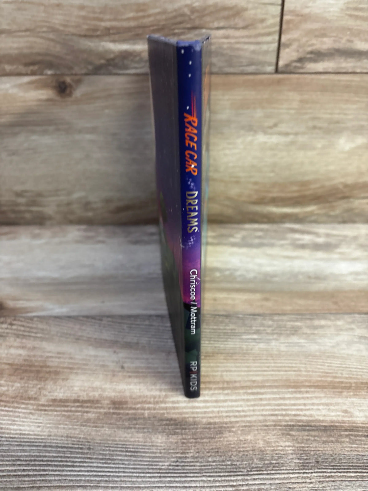 Spine view of Race Car Dreams hardcover book showcasing its blue and purple spine with orange title on a wood-grain background