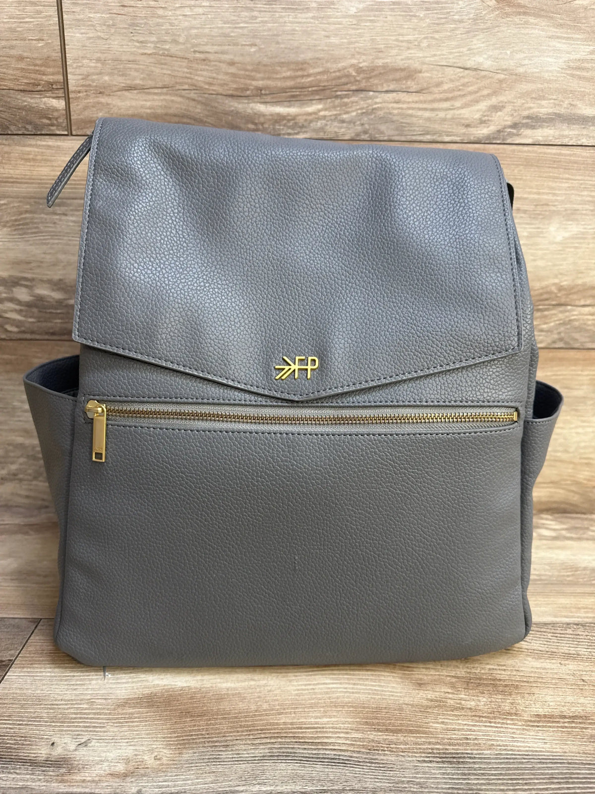 Front view of Freshly Picked Stone Classic Diaper Bag II showing flap-top design, front zip pocket with gold zipper and FP logo on wood background