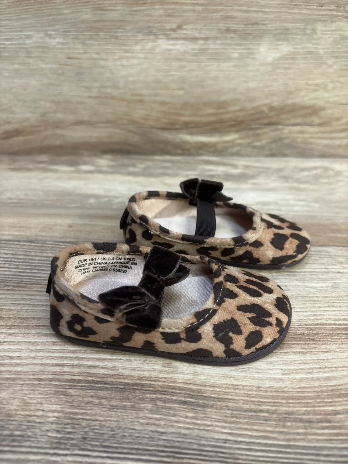 Angled view of H&M leopard-print baby ballet flats size 1/2C showcasing black velvet bows and elastic straps on wood background
