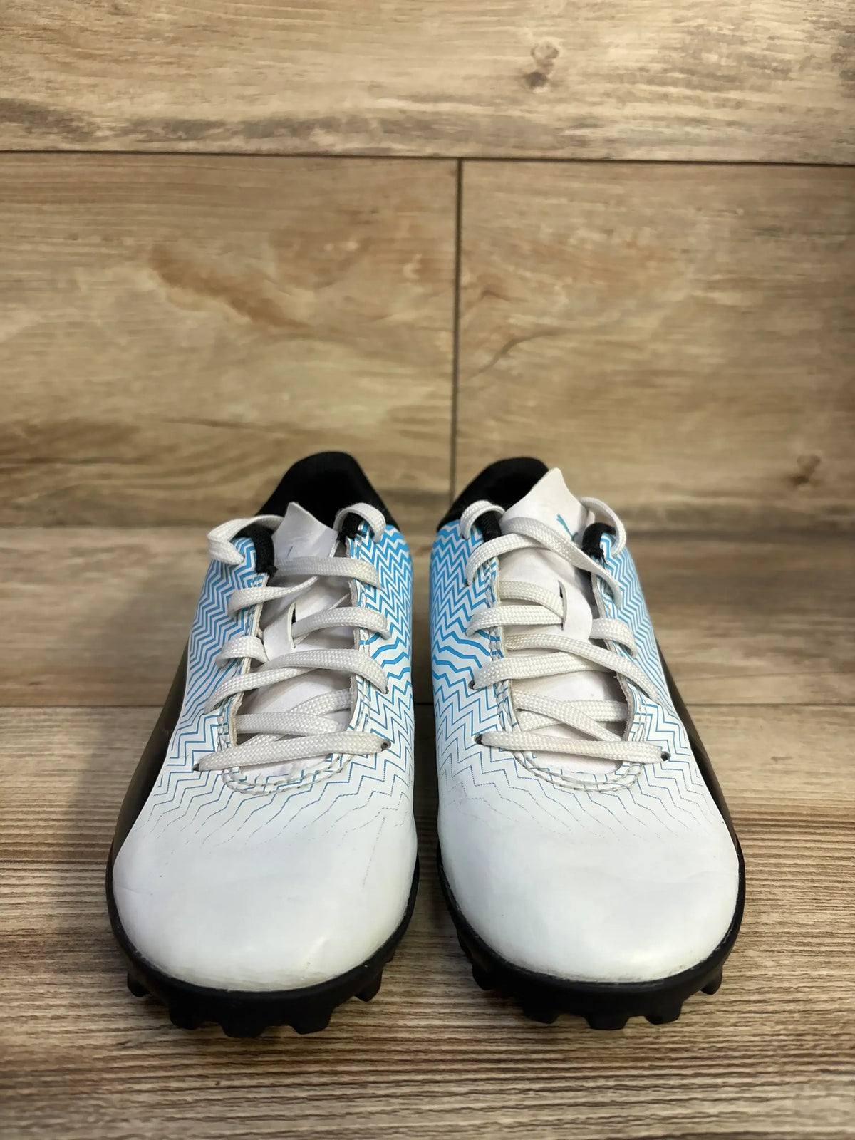 Front view of Puma Rapido II youth soccer cleats showcasing white uppers with blue zigzag pattern, white laces, and black studded outsole on wood floor