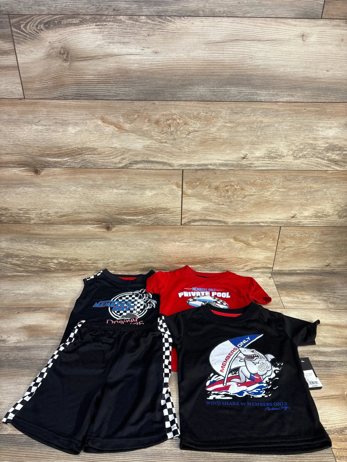 Flat lay of NEW Member’s Only 4pc shirt and short set in black showcasing racing checkered tank shorts and red and navy graphic tees on wood plank floor