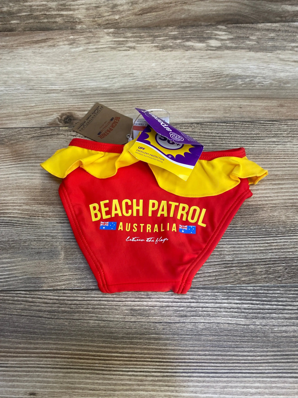 Front view of red toddler swim bottoms with yellow ruffle waist, ‘BEACH PATROL AUSTRALIA’ print on wood plank background