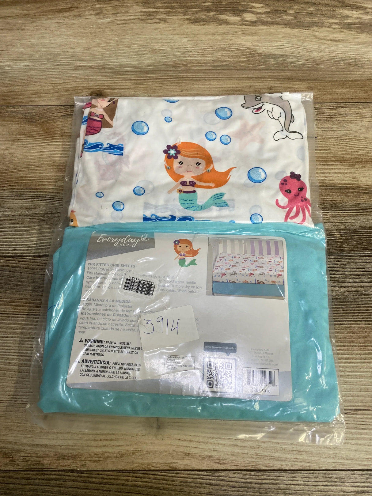 Front view of NEW Everyday Kids 2pk Fitted Crib Sheet packaging with mermaid ocean print and teal solid on wood plank floor