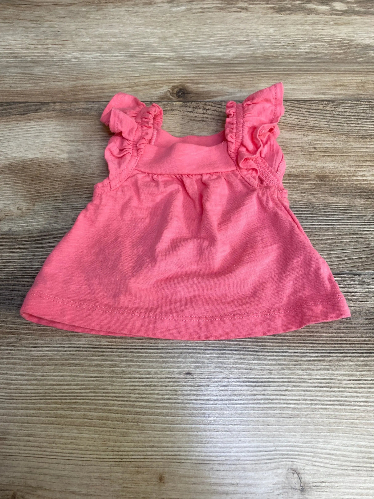 Flat lay of newborn pink Carter’s sleeveless top showcasing ruffled shoulder straps and gathered front on wood background