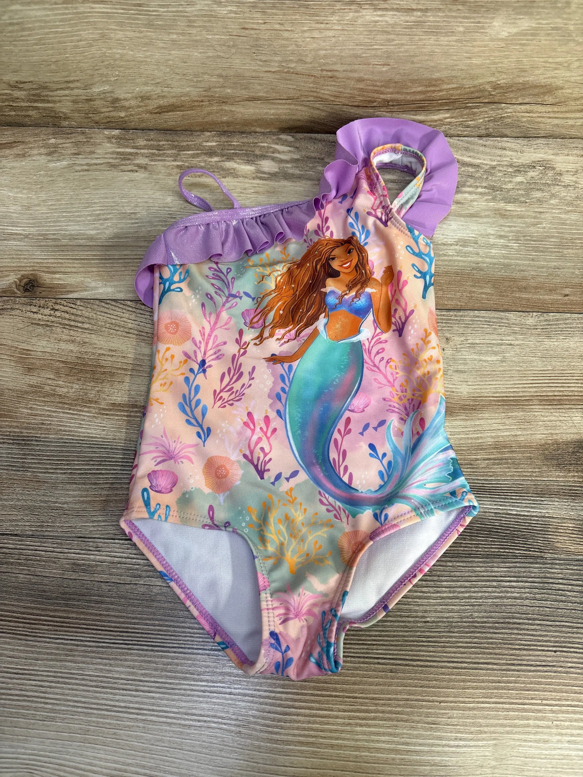 Front view of Disney The Little Mermaid 1pc ruffle swimsuit in size 2T showcasing purple ruffles and Ariel print on wood