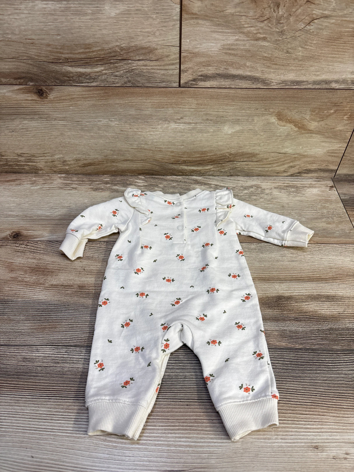 Flat-lay of Carter’s ivory floral coverall showcasing ruffle shoulders, snap-front closure and cuffed legs on wood floor background
