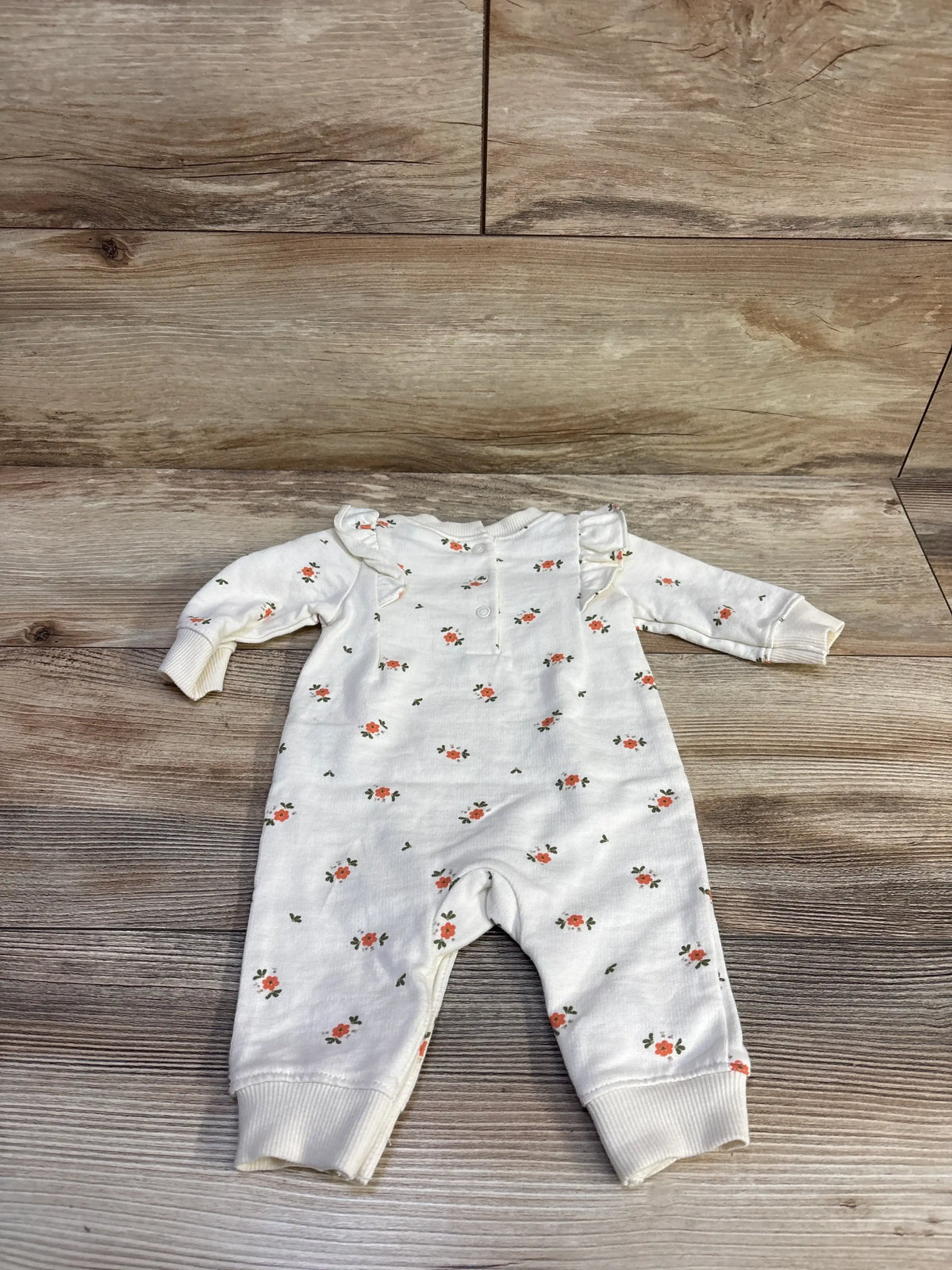 Flat-lay of Carter’s ivory floral coverall showcasing ruffle shoulders, snap-front closure and cuffed legs on wood floor background
